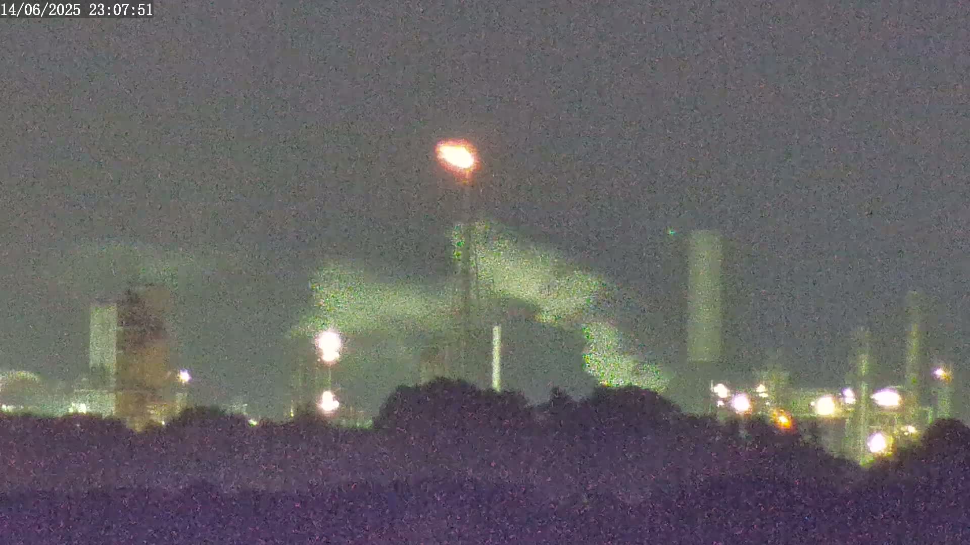 A portion of an ornate clock tower is shown in the foreground, with an industrial complex visible in the hazy distance under a partly cloudy sky.