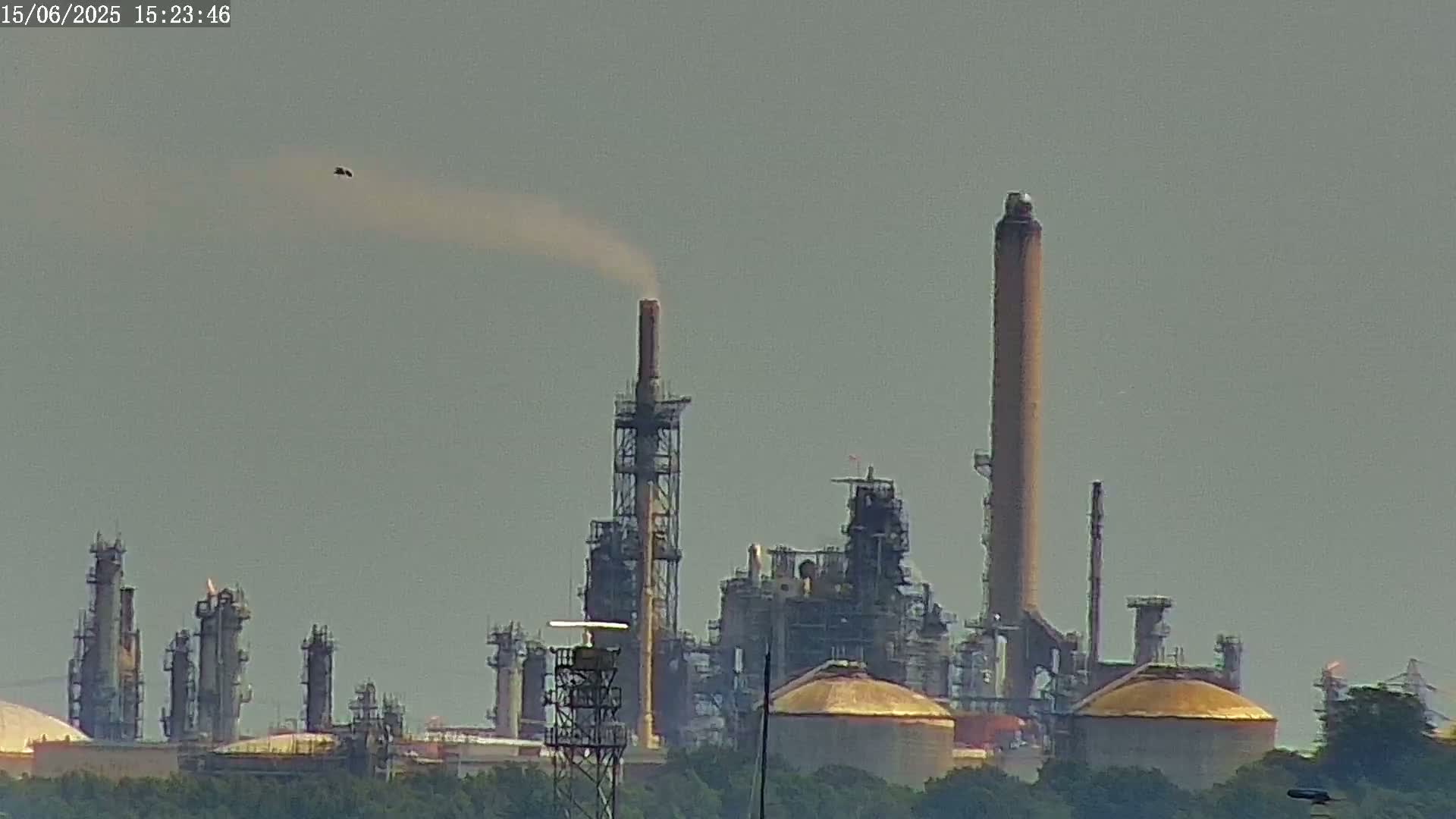 A large industrial plant with several tall smokestacks emits light gray smoke into a hazy sky.