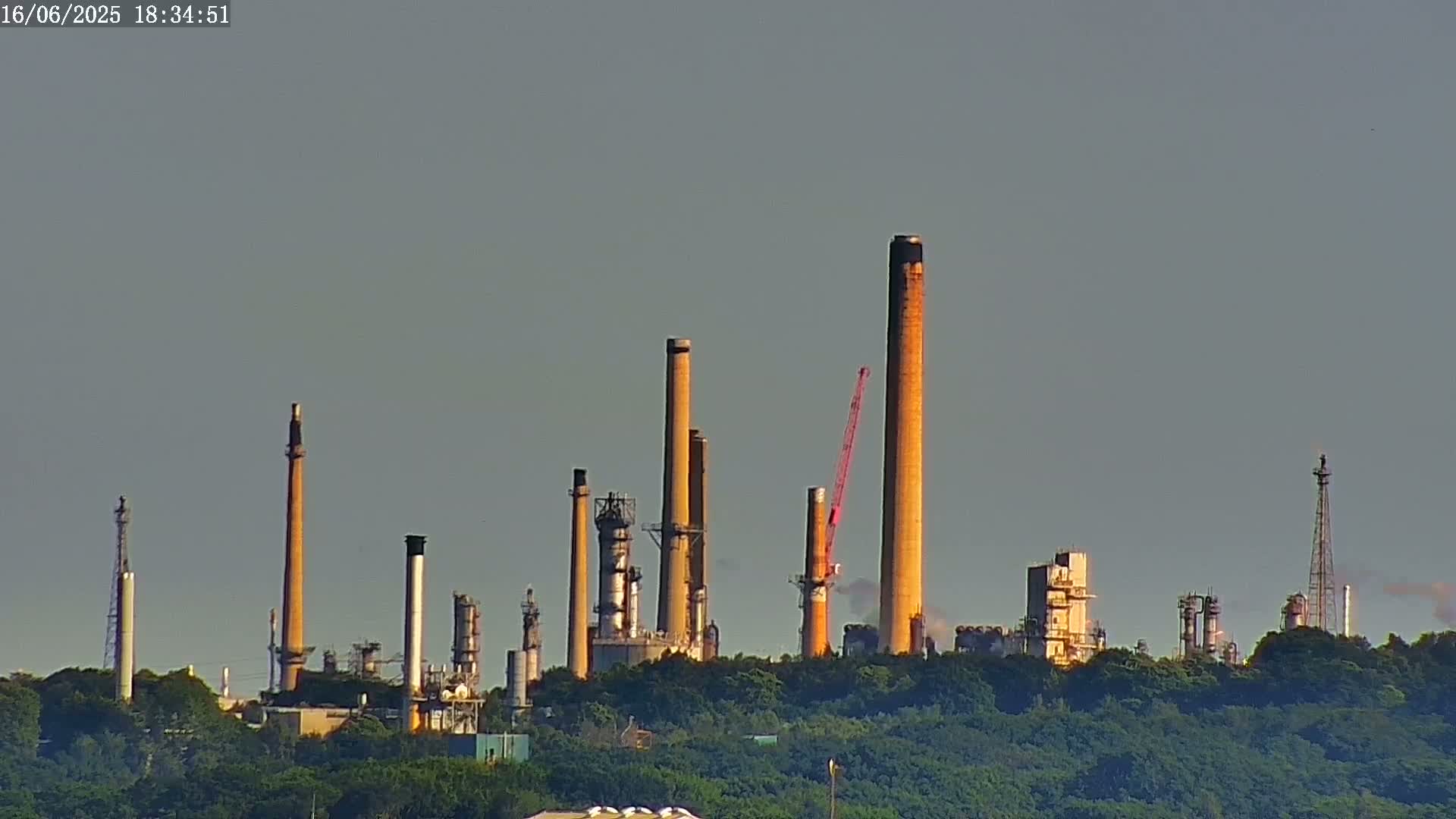 A refinery with numerous tall smokestacks and industrial structures is visible behind a line of green trees under a clear, light gray sky.