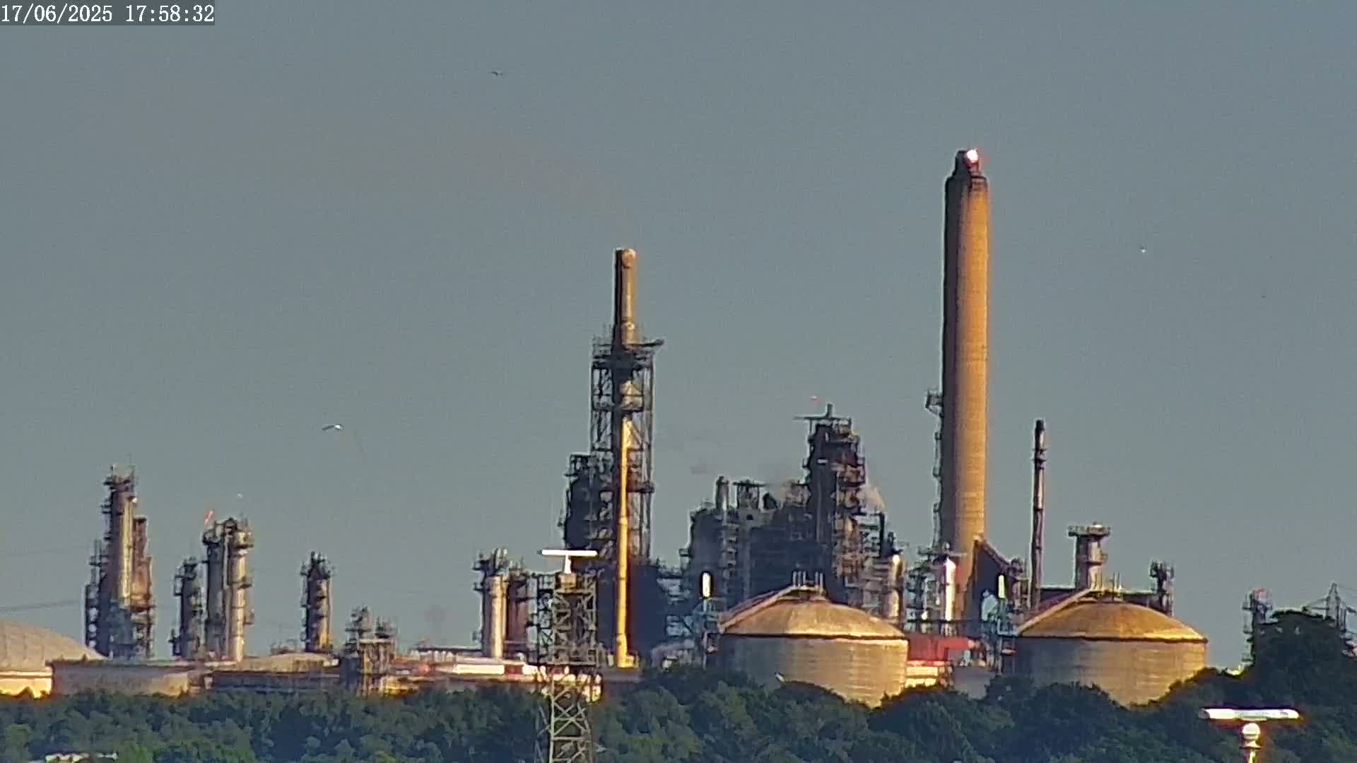 A large industrial complex with tall smokestacks and storage tanks is visible under a clear, light-grey sky.