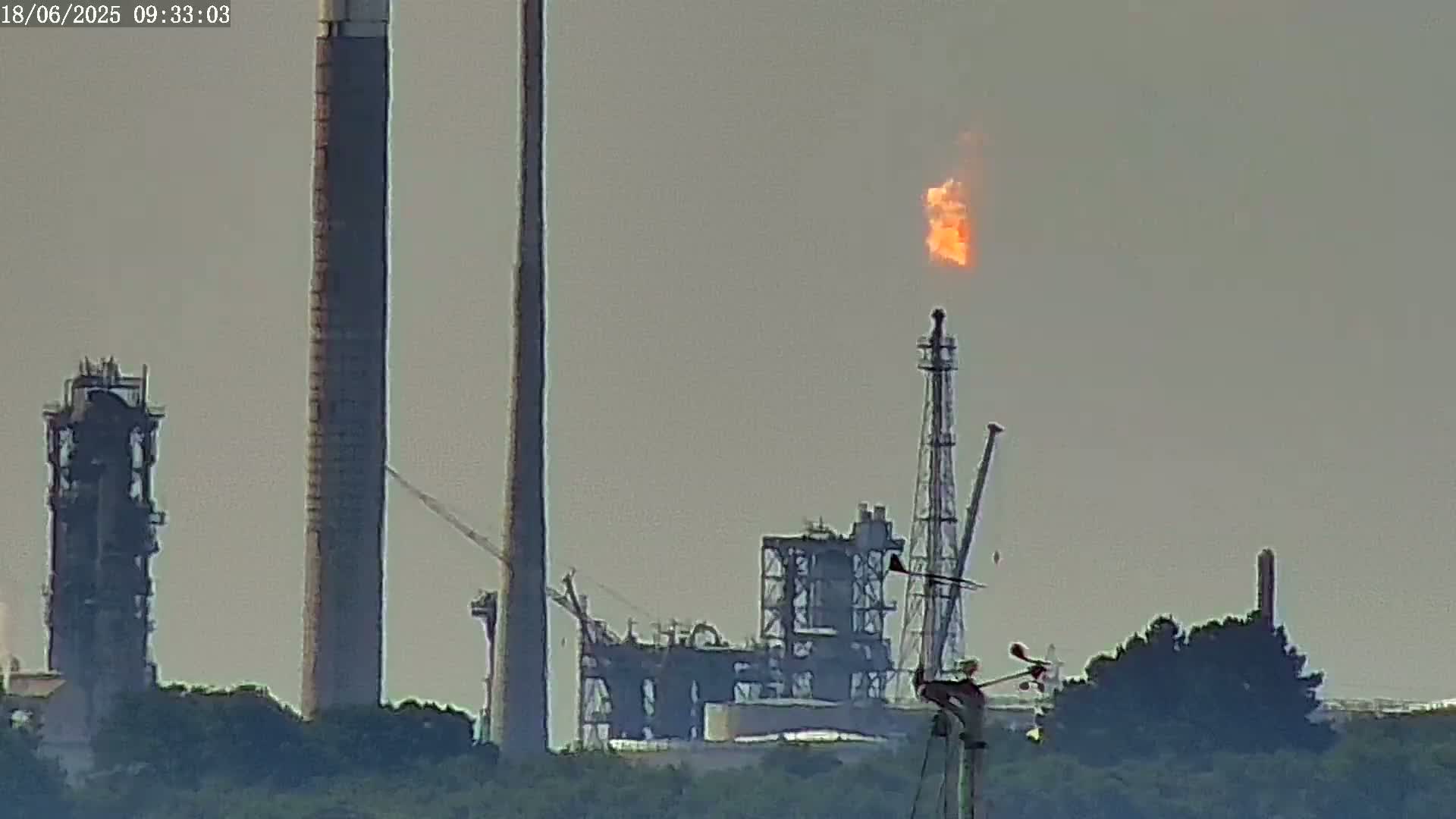 A hazy-looking industrial complex with tall smokestacks, one of which has a bright orange flame burning at its top, is partially obscured by low-lying greenery.