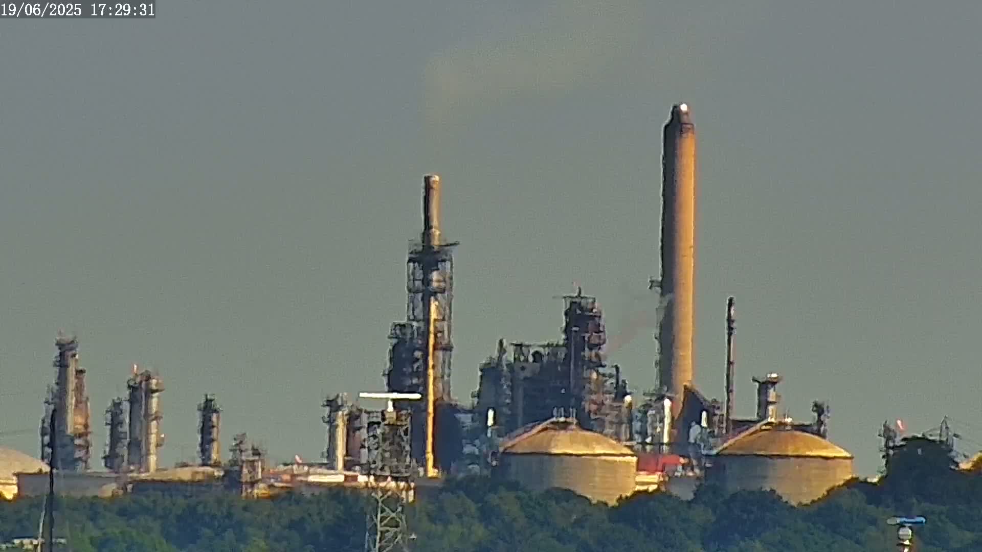 A large industrial refinery complex with numerous tall towers and storage tanks is visible in the hazy distance under a light gray sky.