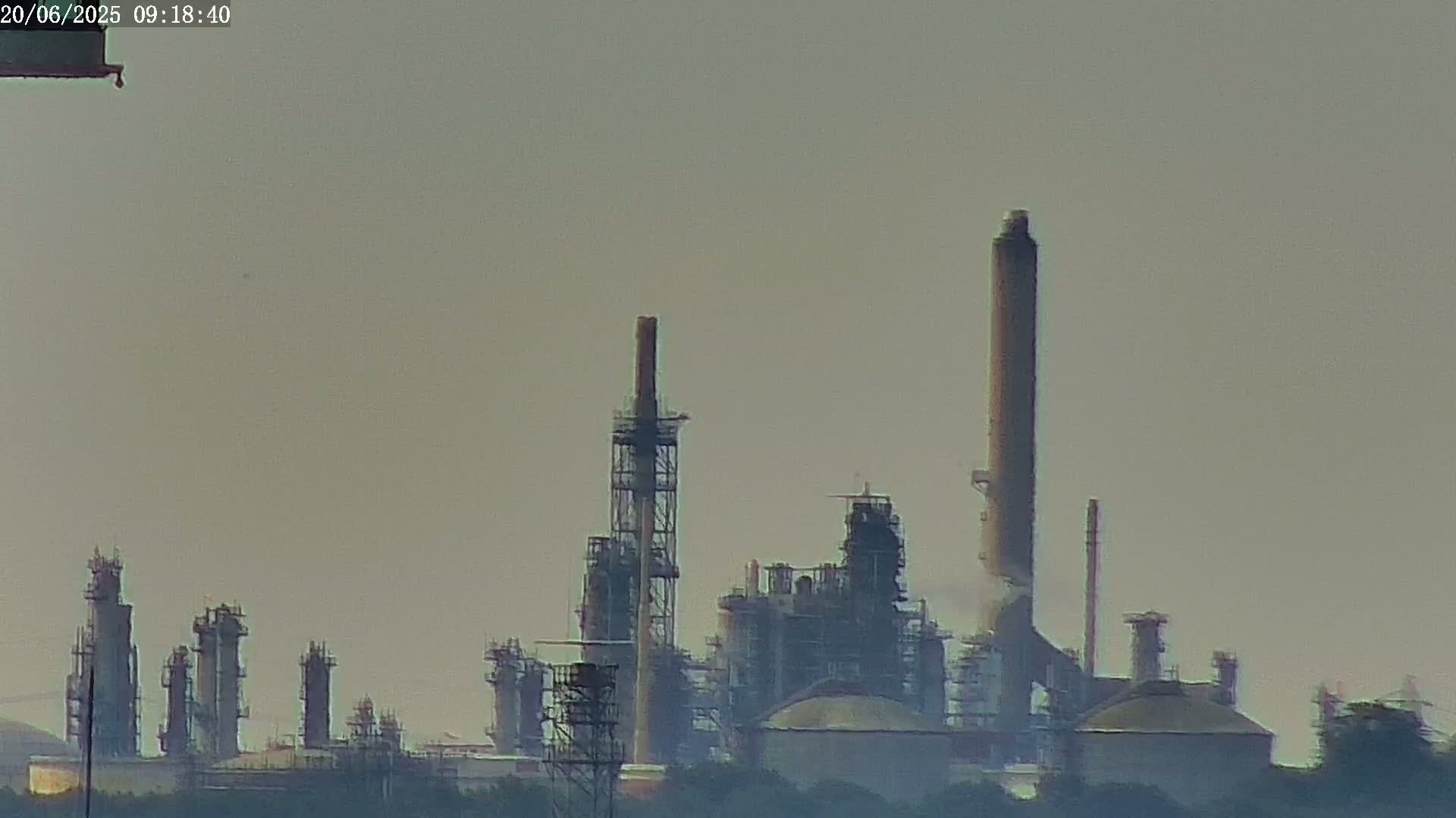 A hazy view of an industrial refinery with tall smokestacks and various structures under a muted sky.