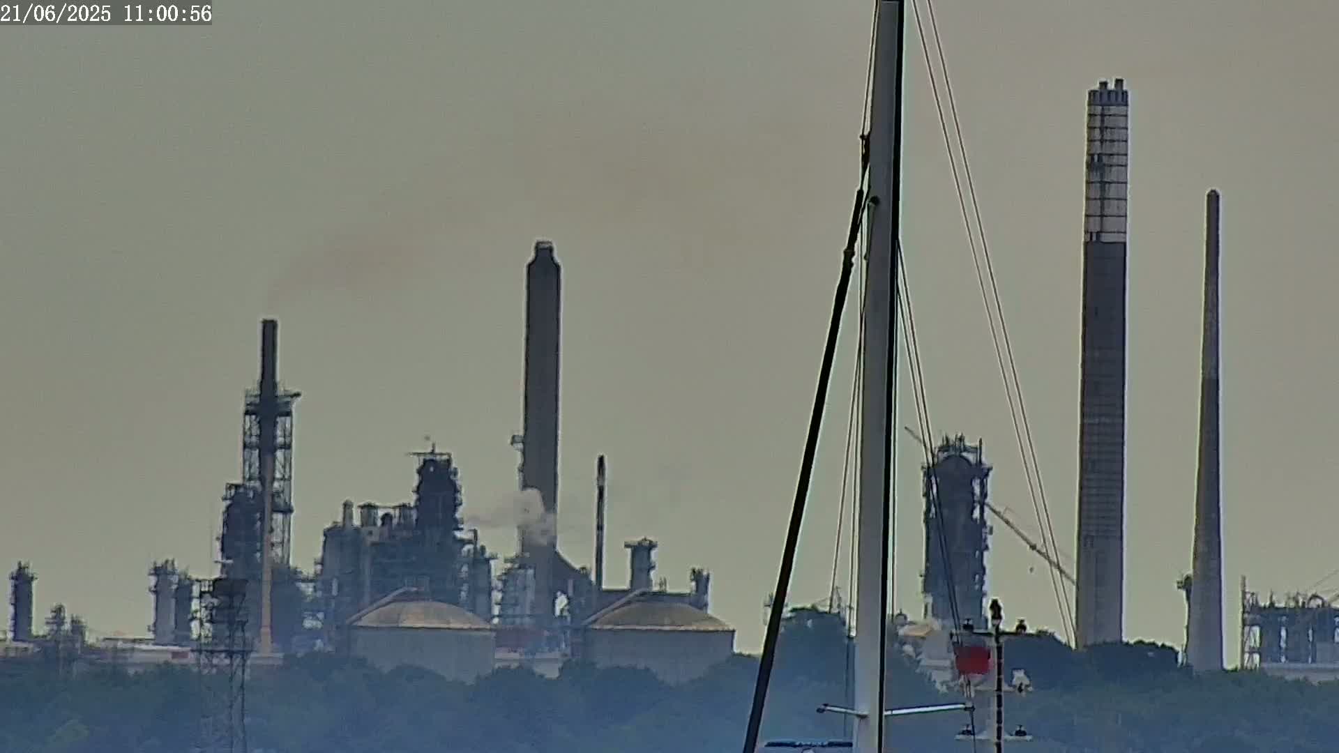 A hazy view of an industrial plant with tall smokestacks is seen from behind the mast of a sailboat.