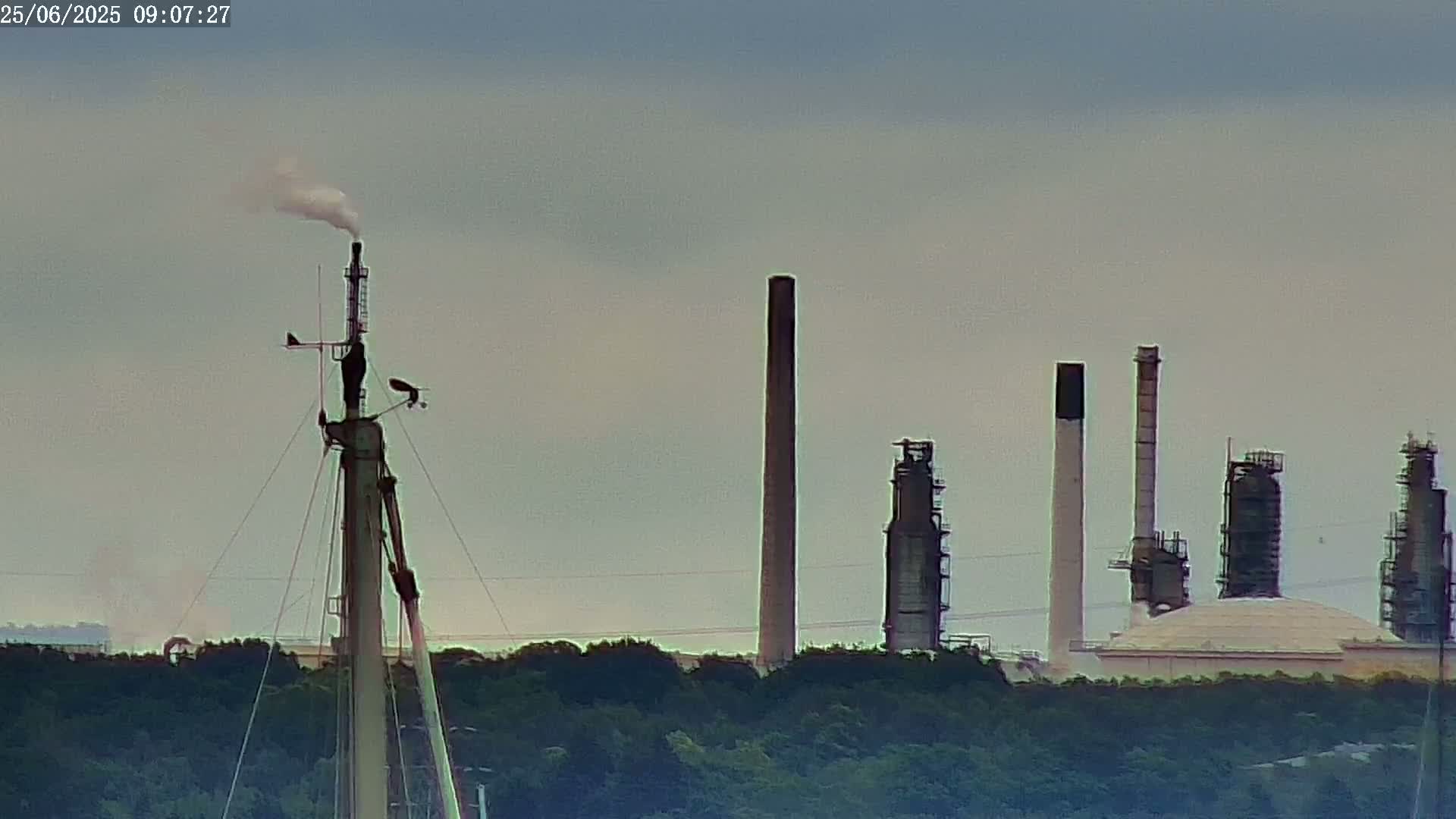 A sailboat mast with a bird perched on it stands in the foreground, with an industrial plant and smokestacks visible in the background under an overcast sky.