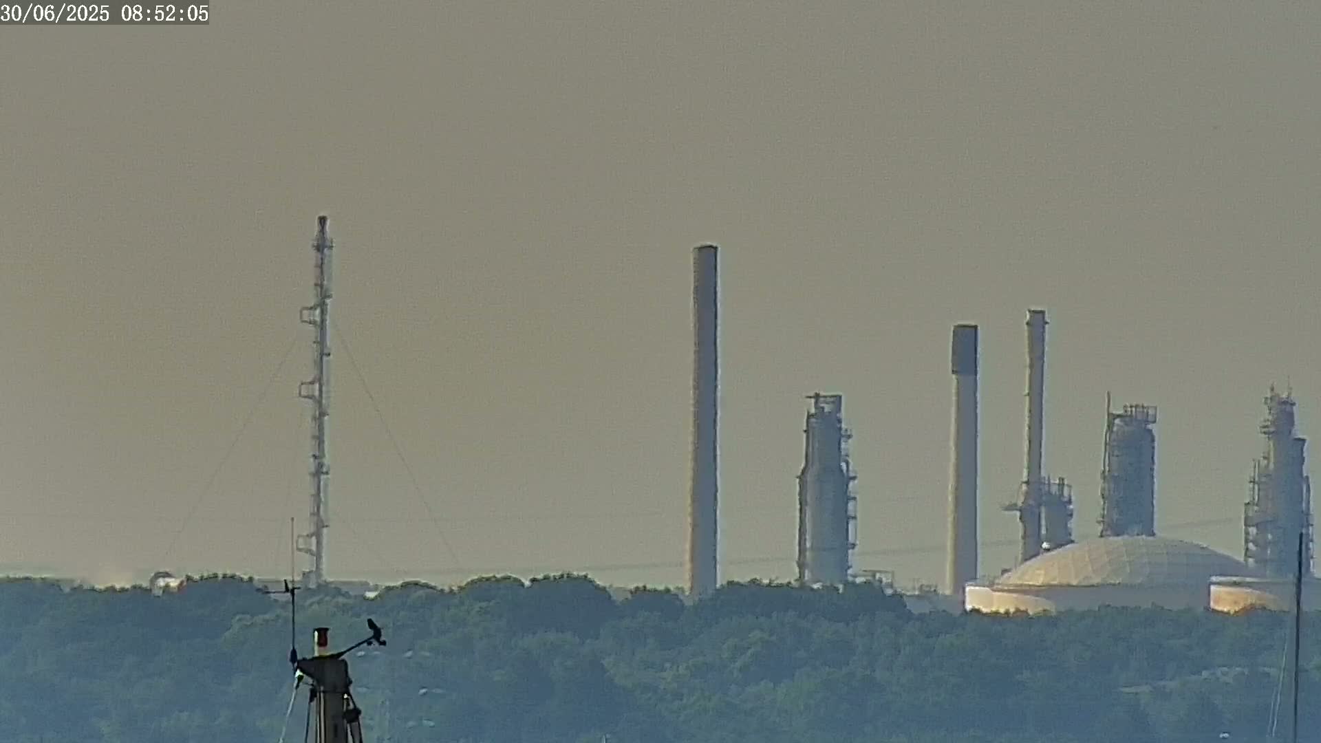 A hazy view shows an industrial plant with several tall smokestacks and a large dome, situated behind a line of green trees.