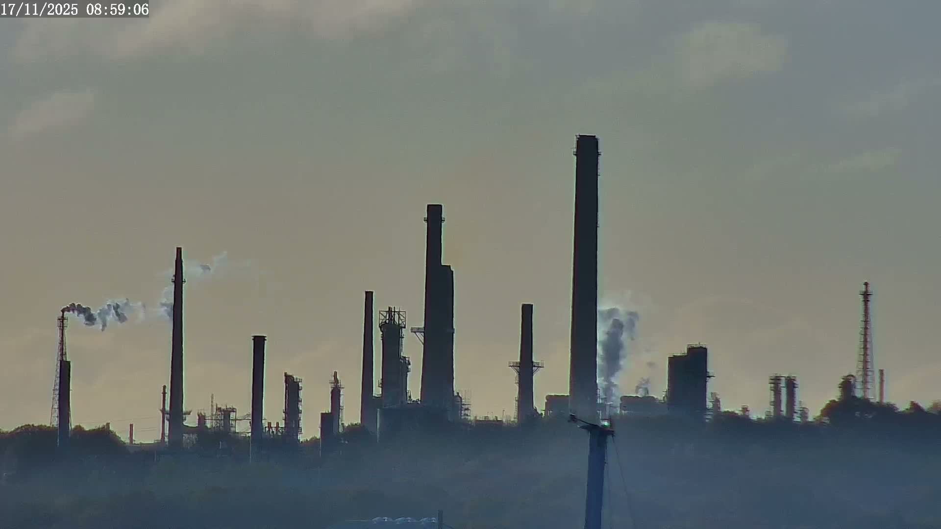 A sprawling industrial refinery with numerous tall smokestacks, some emitting smoke plumes, is partially obscured by dense haze or fog under an overcast sky.
