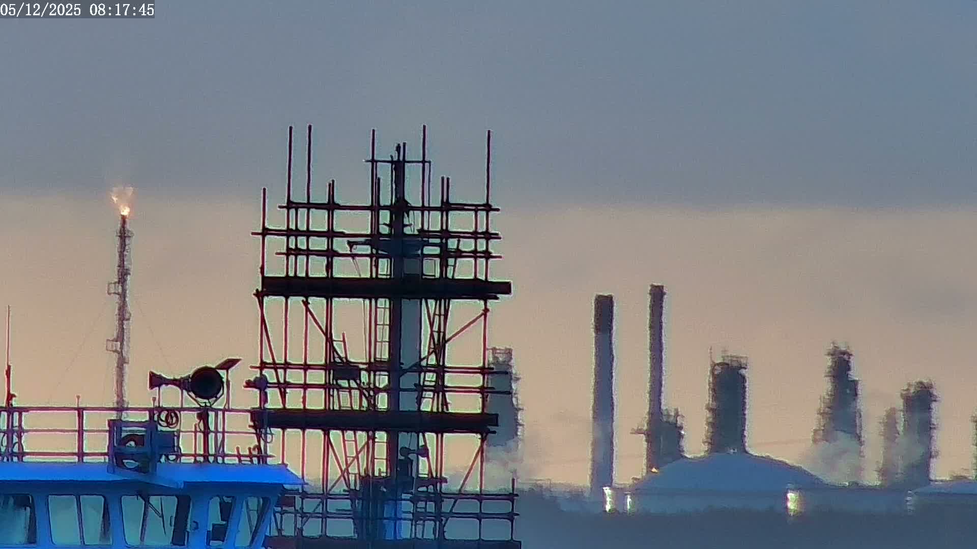 A clear dawn or dusk sky hangs over an industrial scene featuring the top of a blue boat, a flare stack with a bright flame, extensive scaffolding, and numerous factory chimneys emitting plumes of steam.