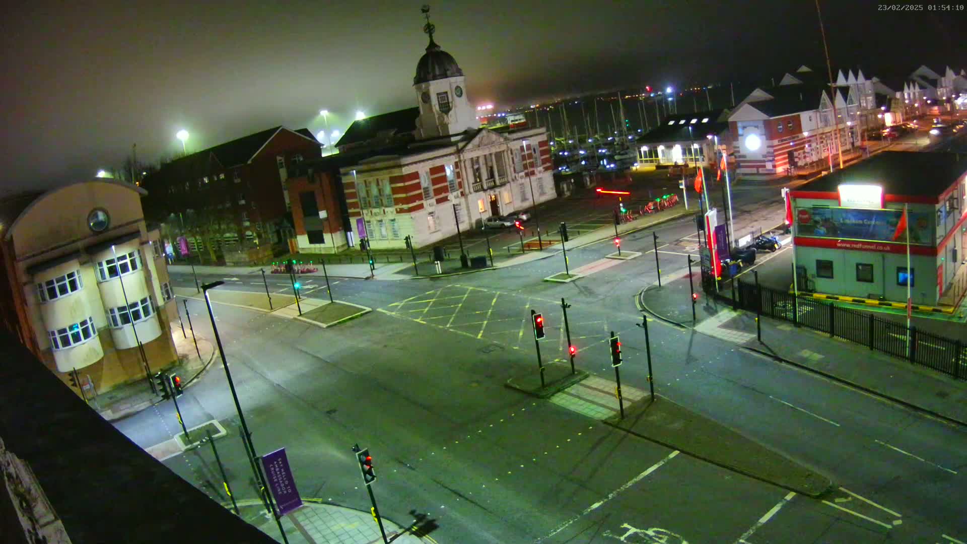 A nighttime, foggy view of a town square with a large building, several smaller buildings, and a marina in the background, all surrounded by empty streets and traffic lights.