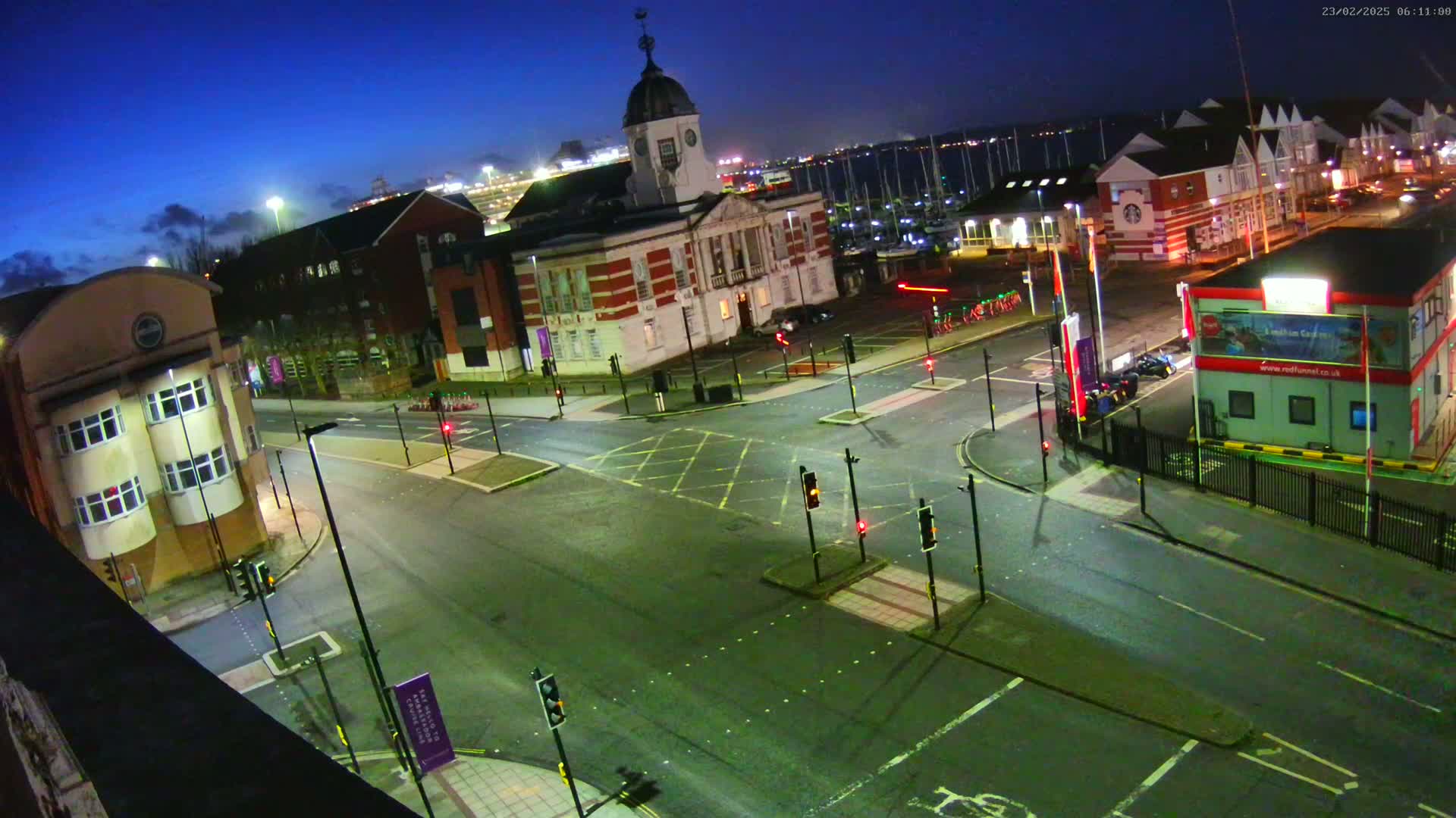An empty road intersection at night, under a dark blue sky, is surrounded by buildings, including a large brick building with a dome, and a harbor is visible in the background.
