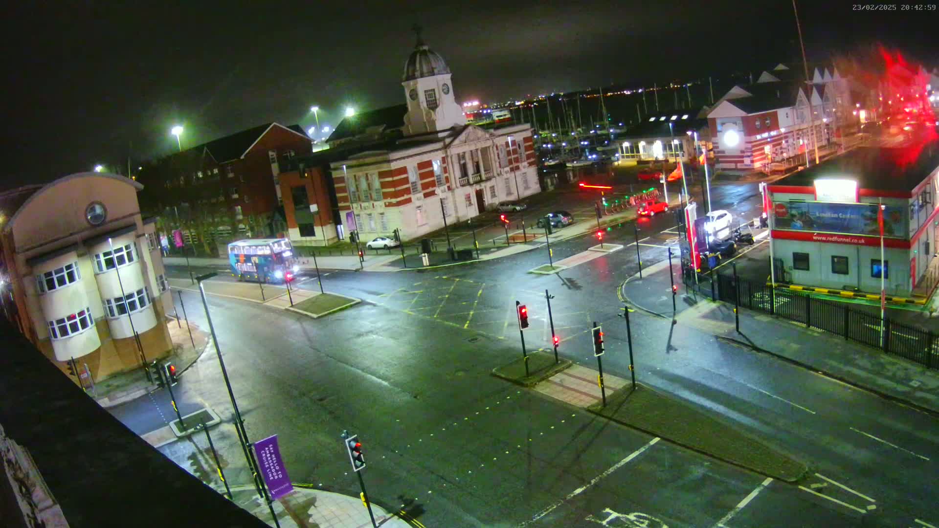 A wet, nighttime street intersection shows several buildings, a bus, and some cars; traffic lights are visible.