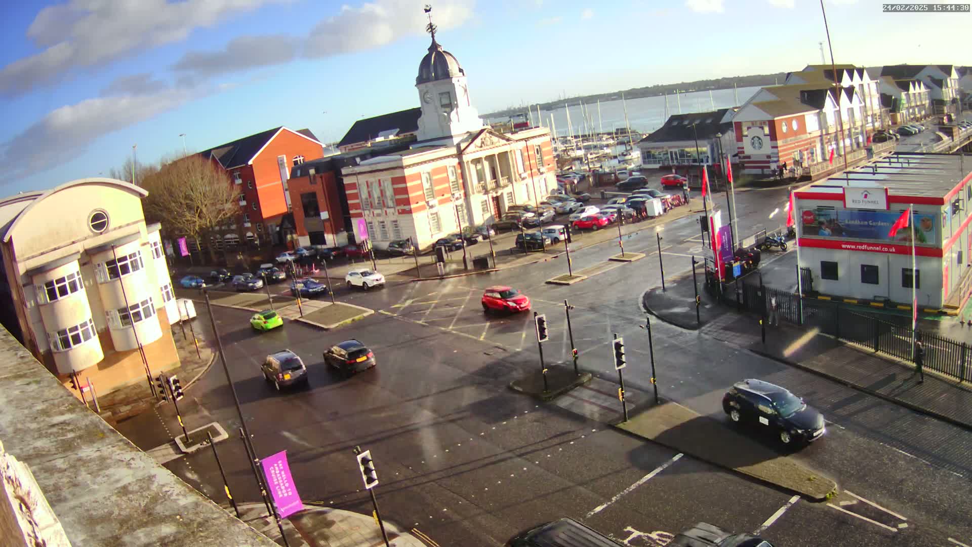 A wet town square with several buildings, including a large brick building with a clock tower, is surrounded by parked and moving cars under a partly sunny sky.