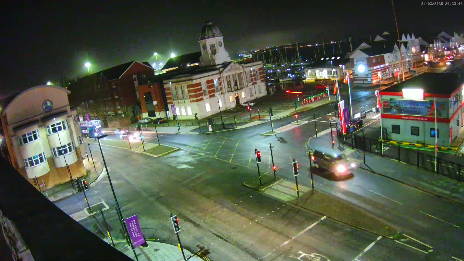 A wet, nighttime street intersection shows several vehicles, including a bus, at a crossroads near a large brick building and a marina.