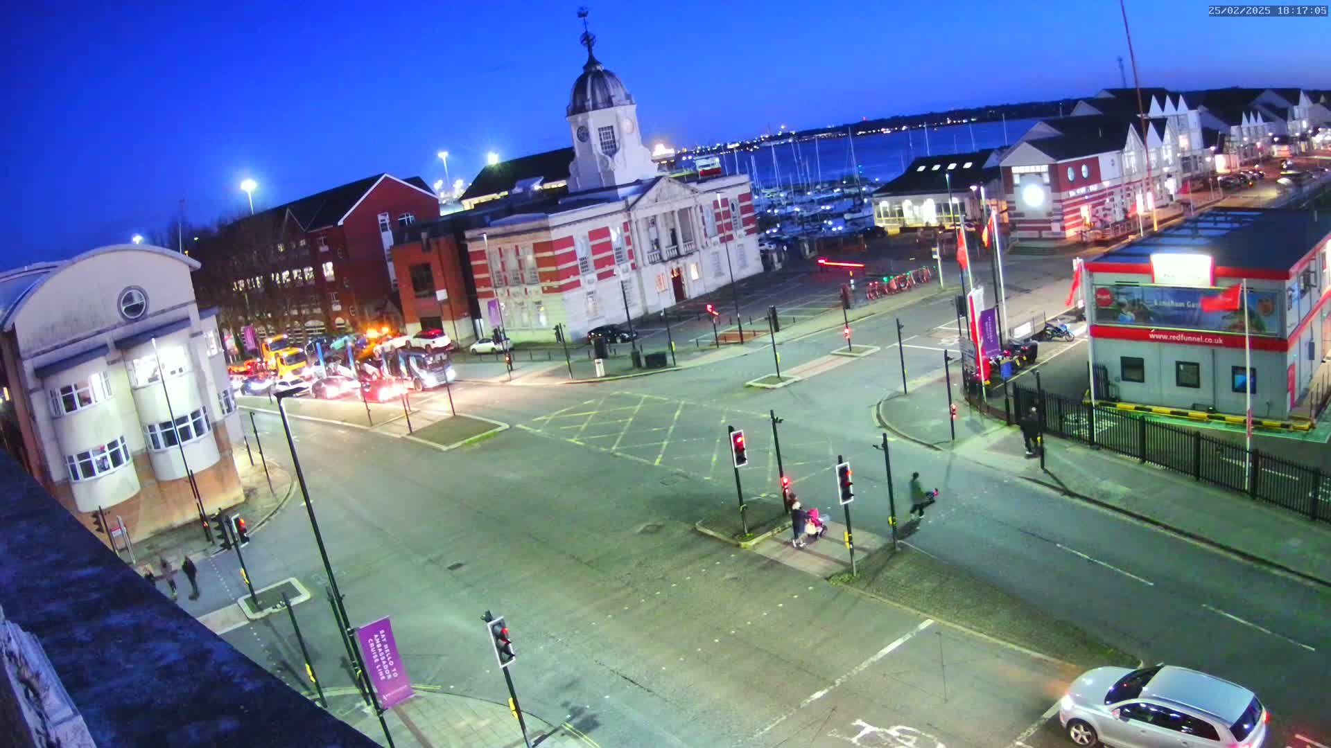 A twilight view of a harborside town square with several buildings, vehicles, and pedestrians at a crosswalk, under a clear blue sky.