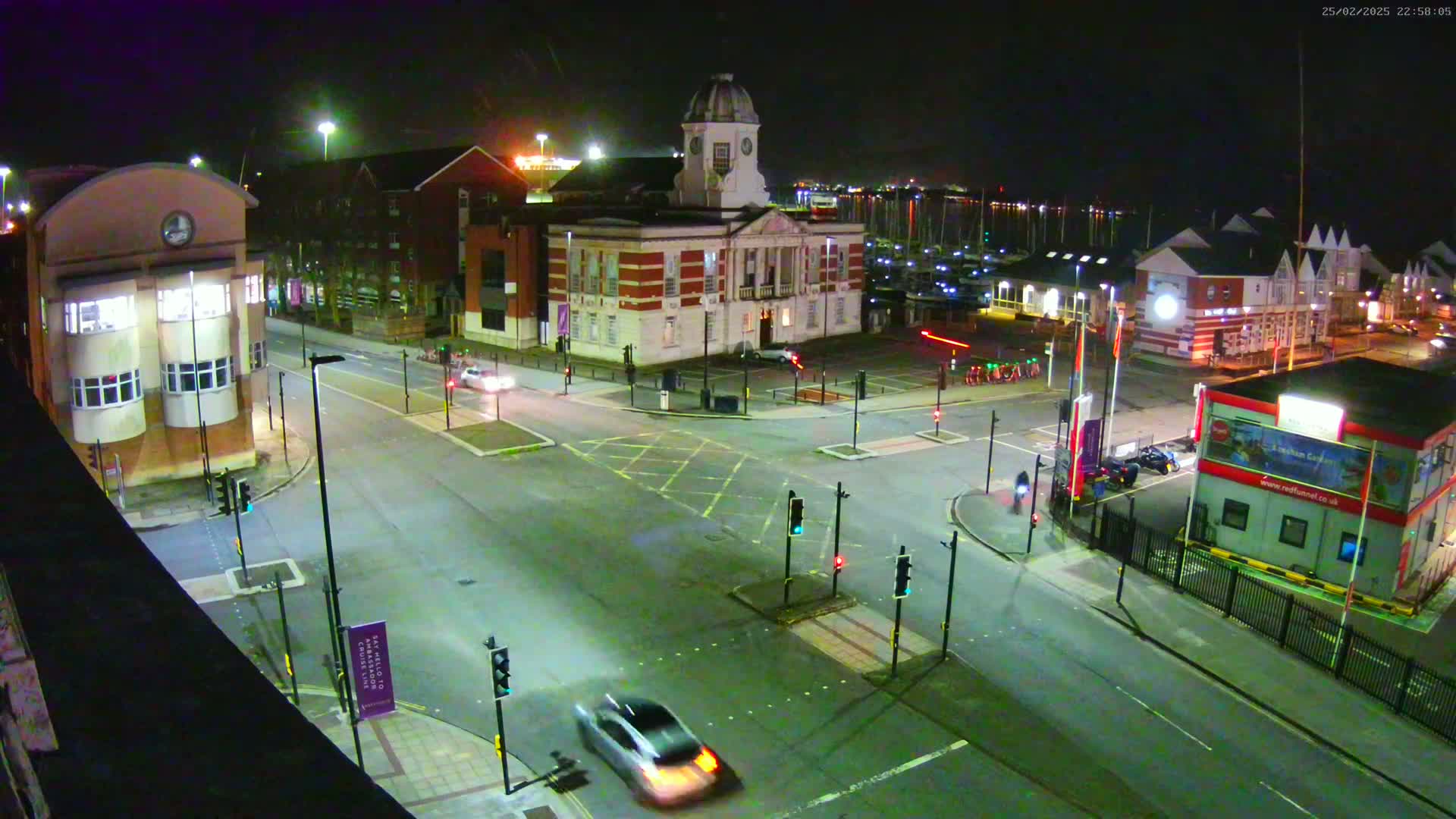 A nighttime, overhead view of a city intersection with several buildings, including a large brick building with a clock tower, illuminated by streetlights and surrounded by cars and traffic signals.