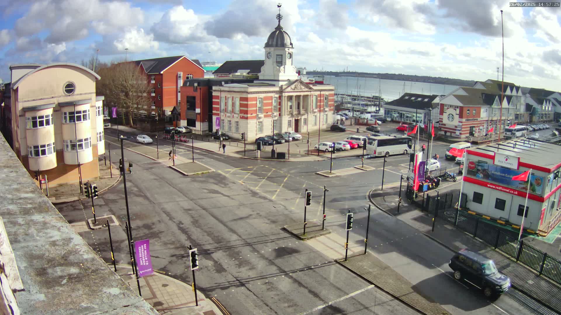 A high-angle view of a town square on a partly sunny day, featuring a brick clock tower building, surrounding buildings, parked cars, and a busy intersection with traffic lights.