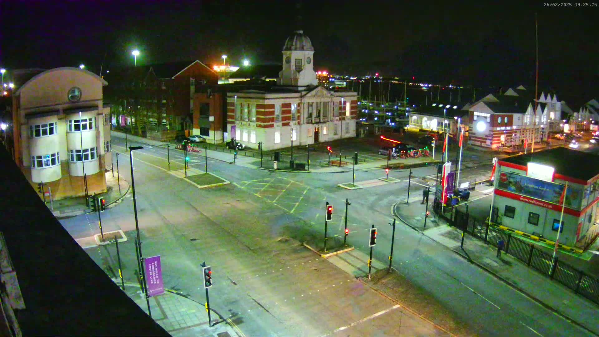 An empty nighttime street intersection with several buildings, including a clock tower, and a marina in the background under clear skies.