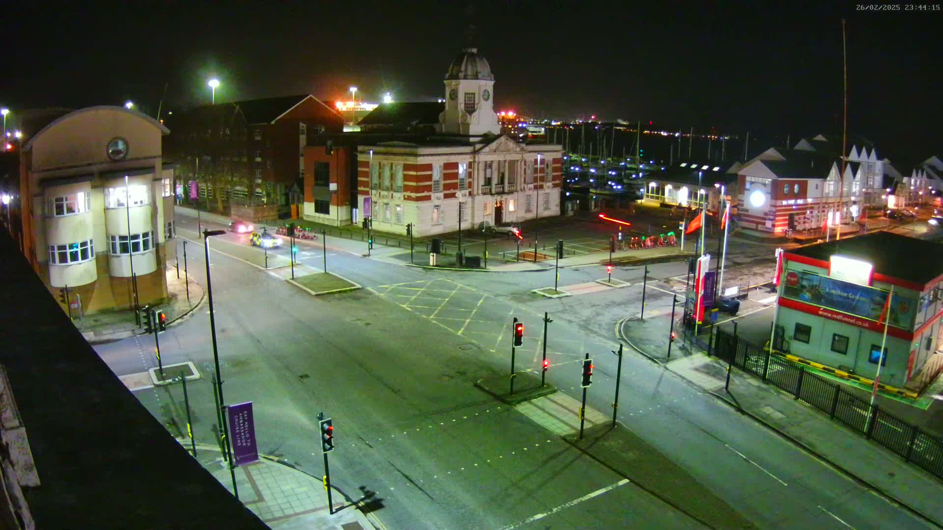 A nighttime, high-angle view of a street intersection near a harbor, with several buildings including a clock tower and traffic lights.