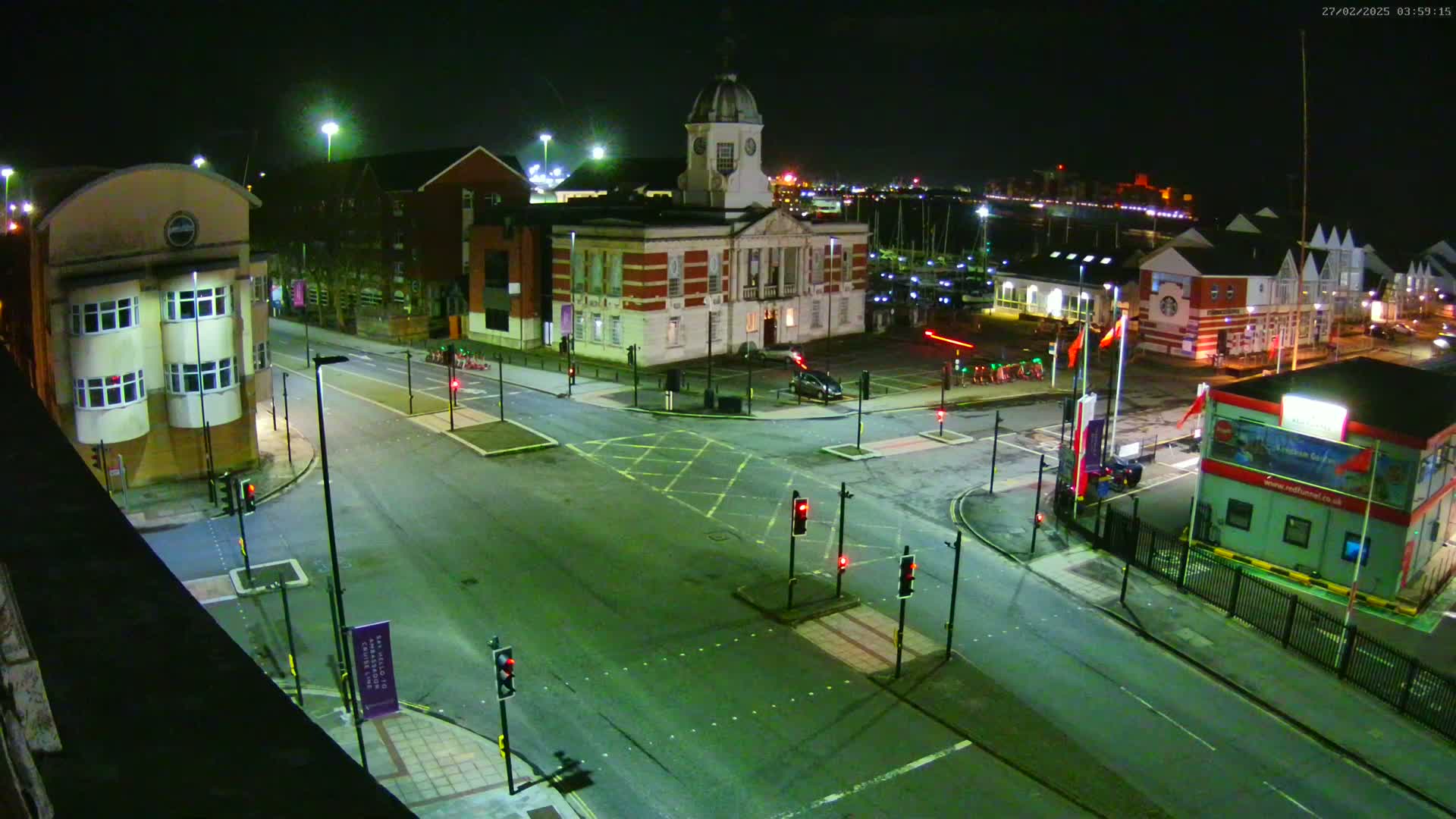 A nighttime aerial view of a town square with a large brick building, several smaller buildings, and a roadway intersection with traffic signals.