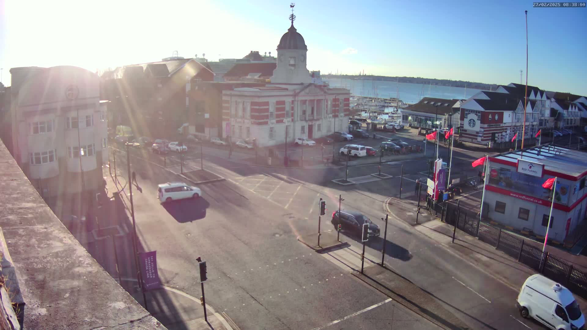 A sunny day overlooking a town square with a large building and several cars at a crossroads near a waterfront with boats.