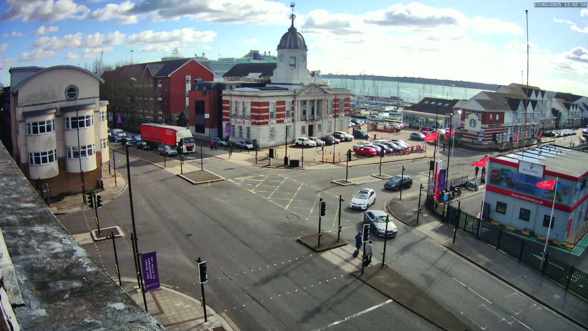 A partly sunny day shows a street intersection with several cars and pedestrians near a brick building with a dome,  adjacent to a marina with boats.