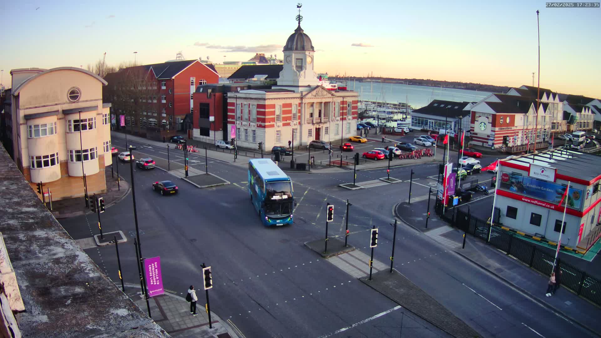 A busy intersection with several cars and a bus, near a body of water under a partly cloudy evening sky.