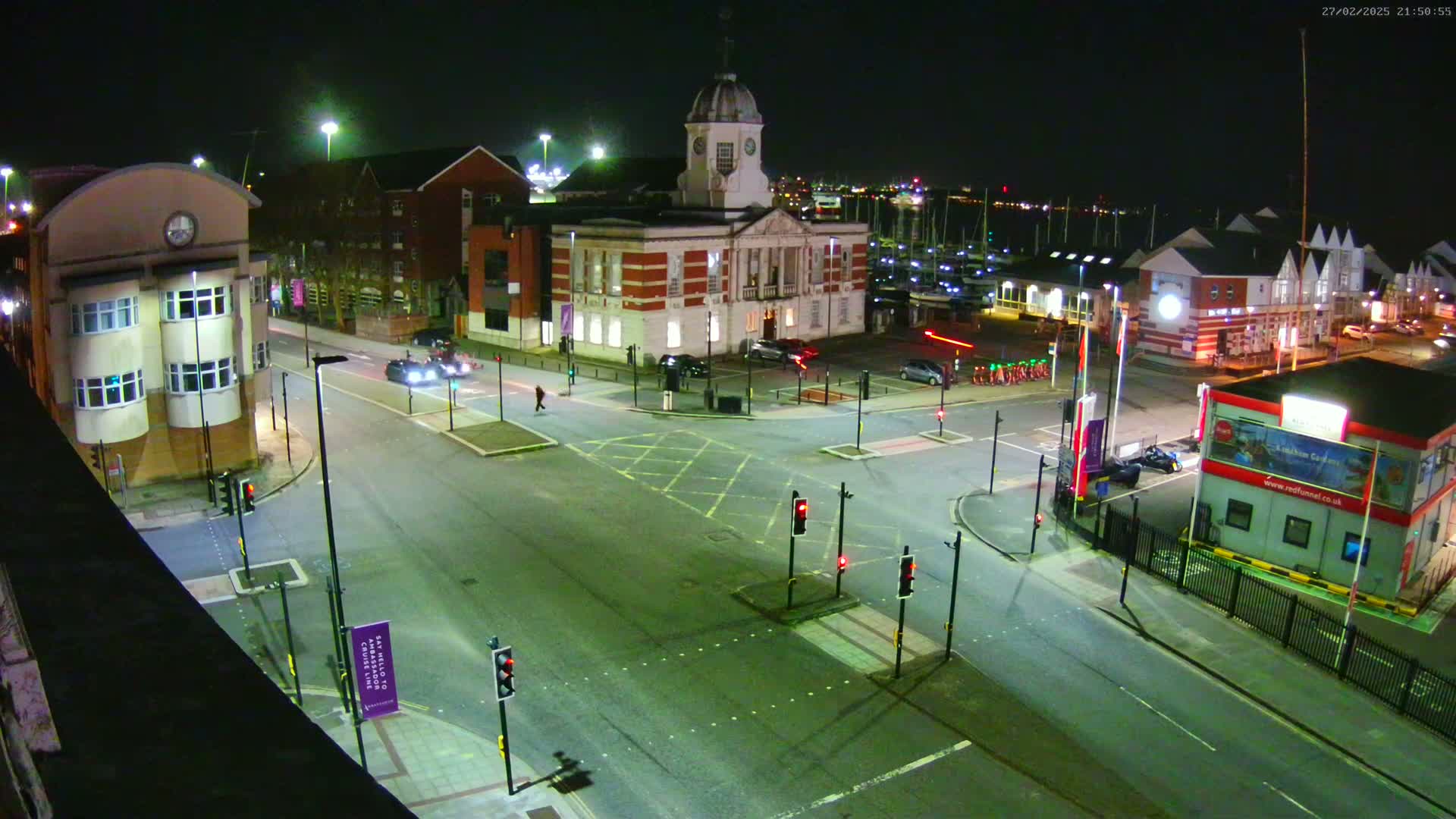 A nighttime aerial view of a town square intersection with several buildings, including a clock tower, and cars at a standstill at traffic lights.