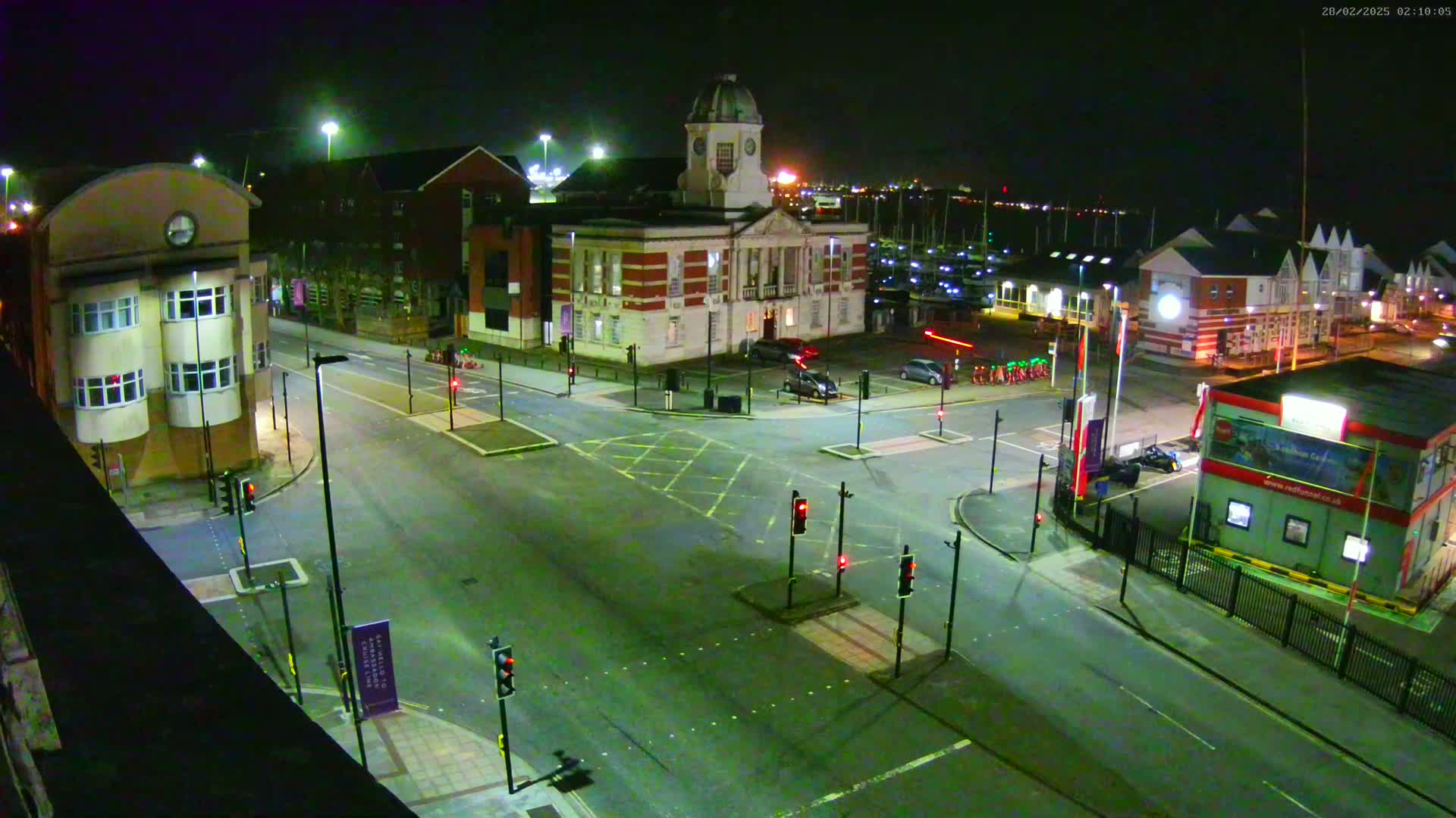 A nighttime aerial view of a town square with a clock tower building at its center, surrounded by other buildings, roads, and traffic lights, under dark, clear skies.