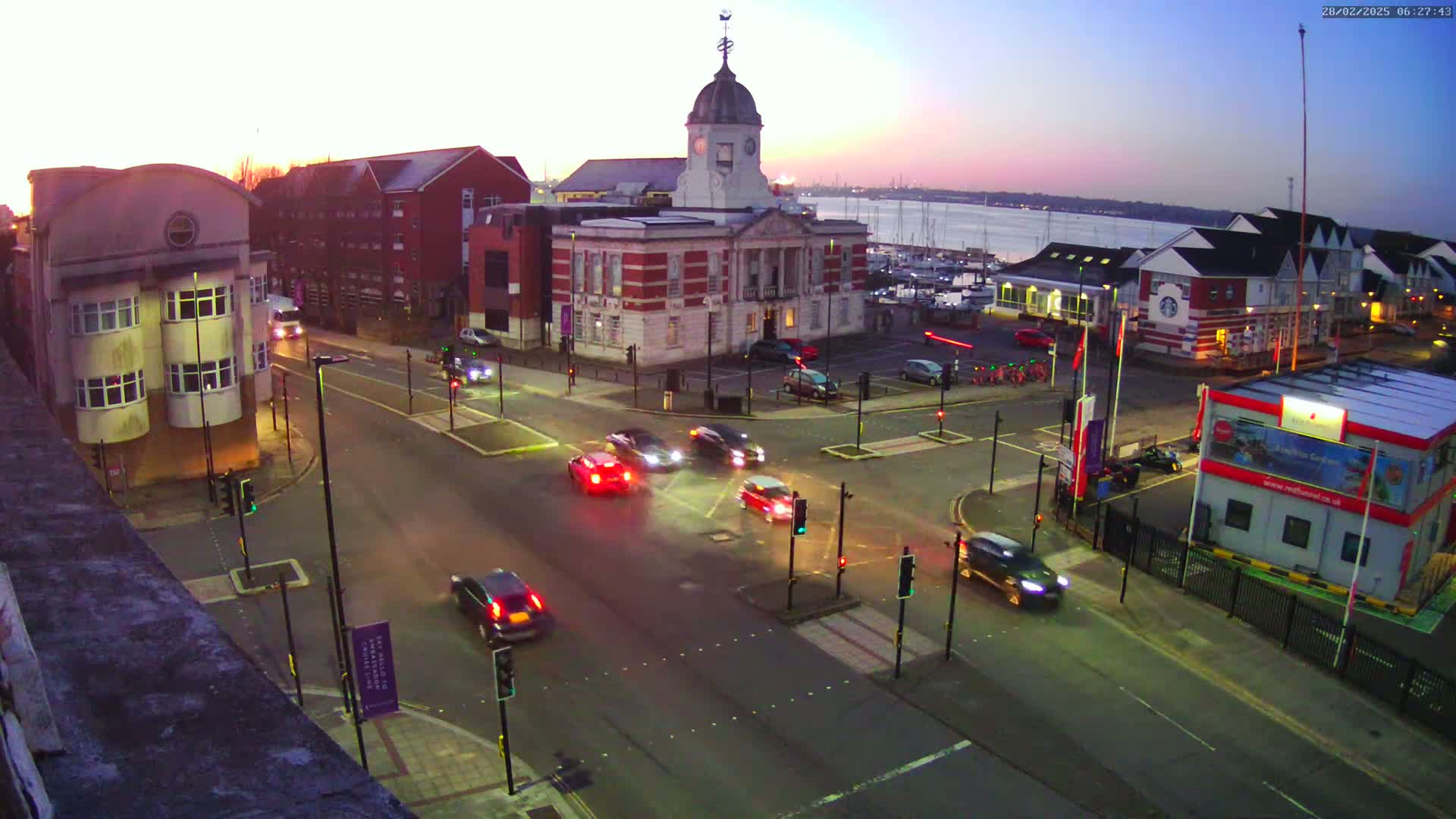 Several cars are driving through a city intersection at twilight, near a waterfront with boats, under a mostly clear sky.