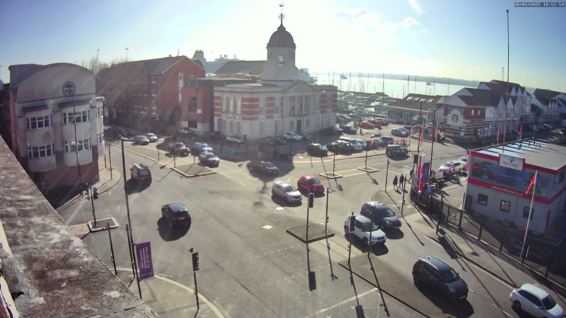 A sunny day reveals a town square with a brick building at its center, surrounded by parked cars and traffic, with a body of water and boats visible in the background.
