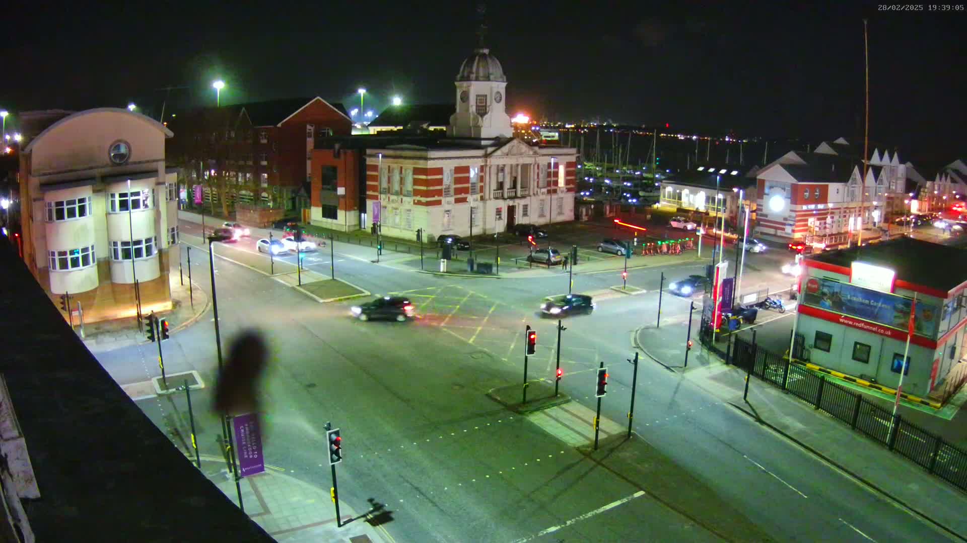 A nighttime, high-angle view of a town square intersection with several cars at a traffic light, surrounded by buildings including a clock tower and marina in the background.