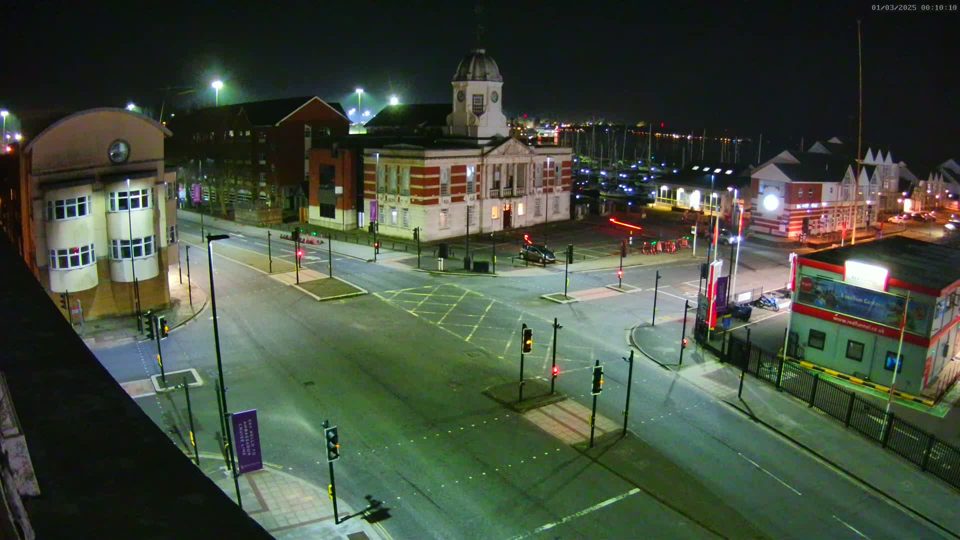A nighttime aerial view of a street intersection near a harbor, with several buildings, including a clock tower, and traffic lights.