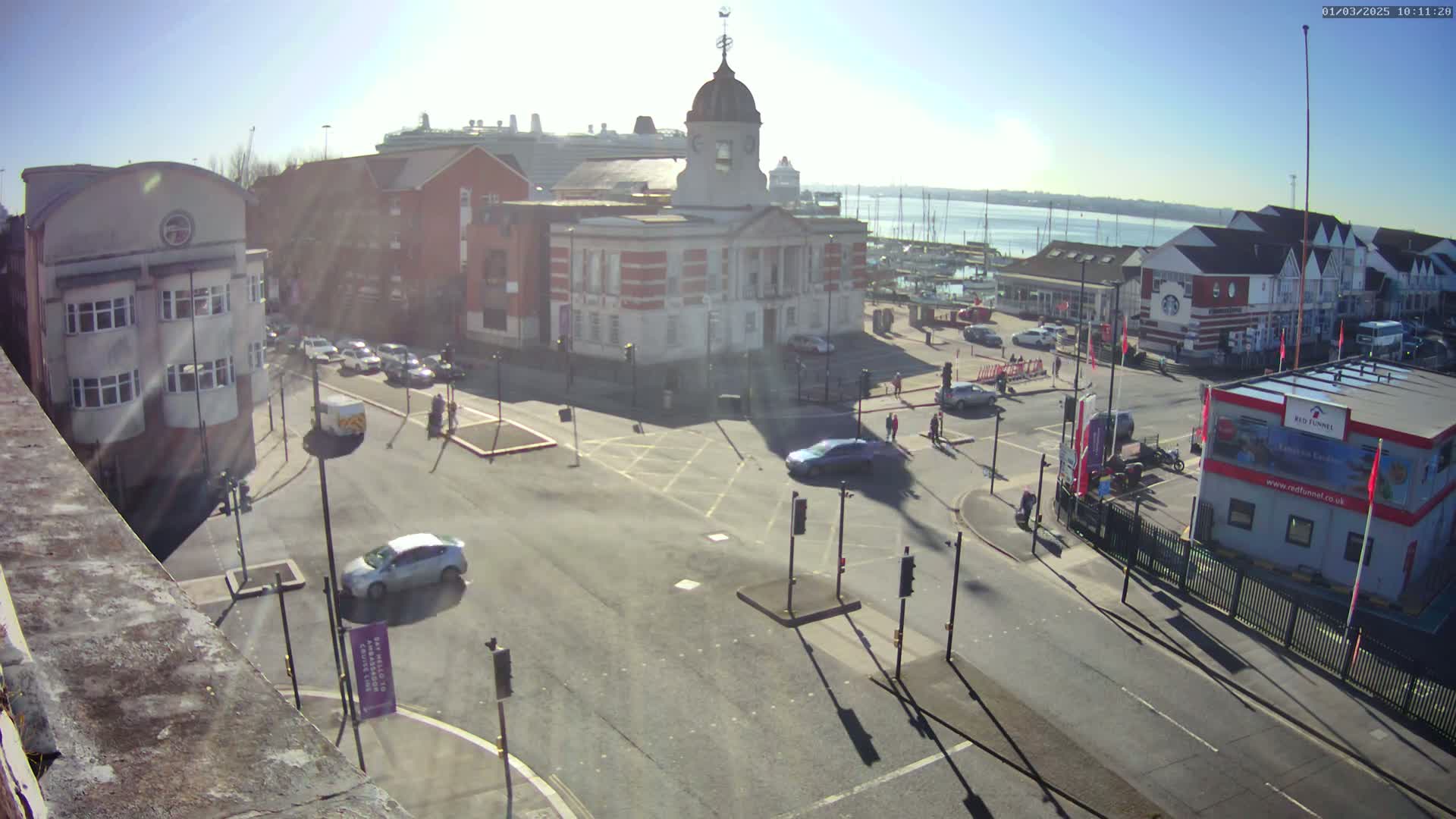 A sunny day reveals a city square with a large building, several cars, and a harbor with boats and a cruise ship in the distance.
