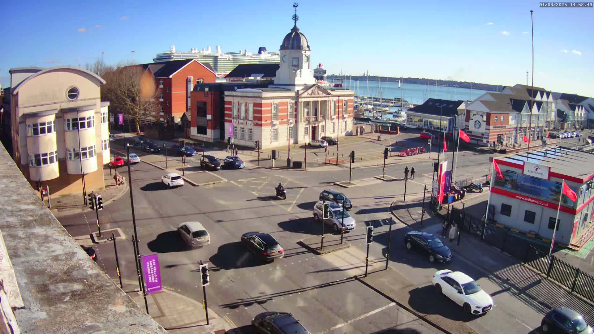 A sunny day shows a busy intersection with several cars and a motorcyclist, near a waterfront area with docked boats and a large cruise ship in the distance,  with a brick building in the center of the image.