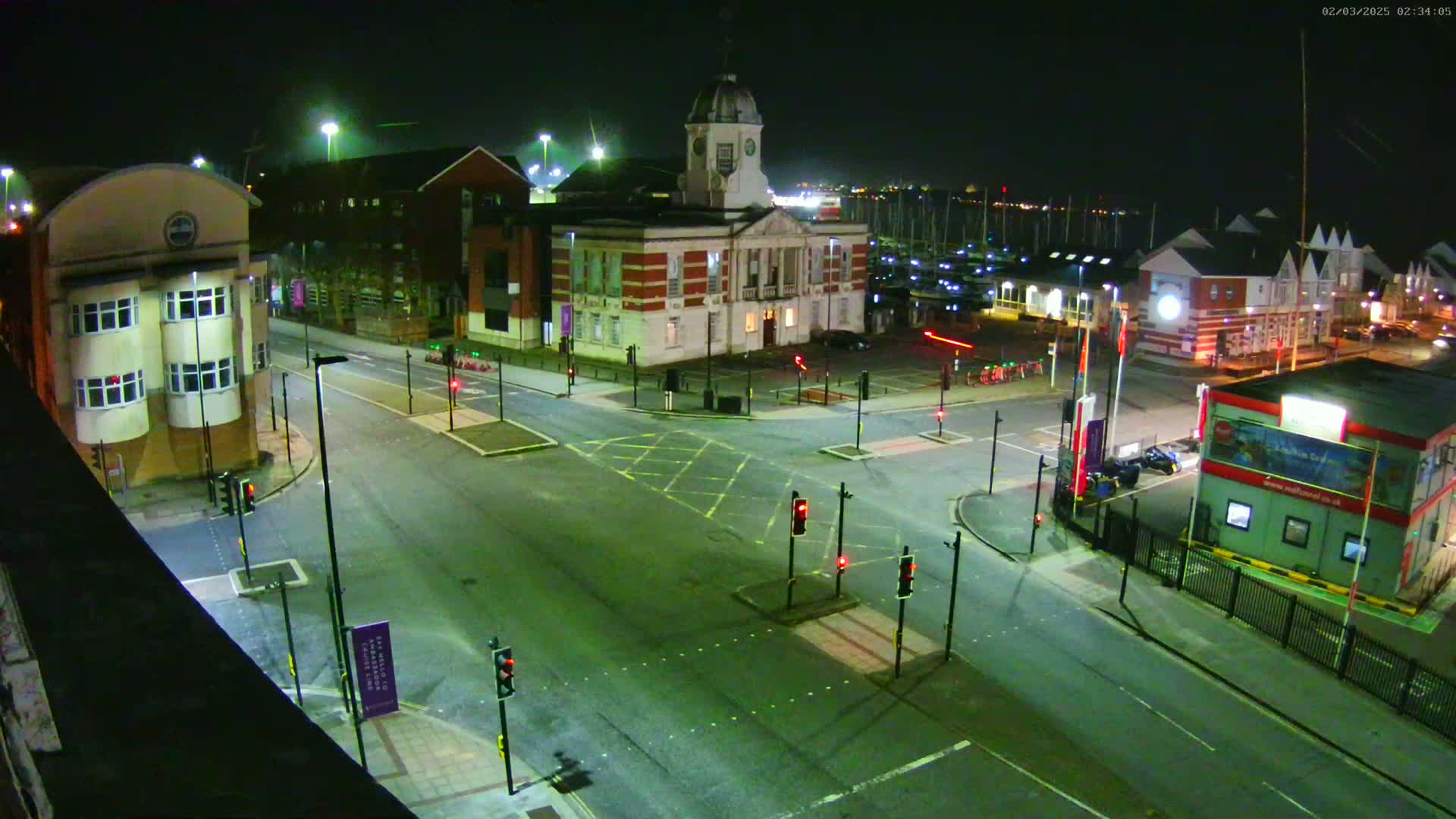 A nighttime aerial view of a town square with a clock tower building, several other buildings, a marina in the background, and traffic signals at a crossroads.