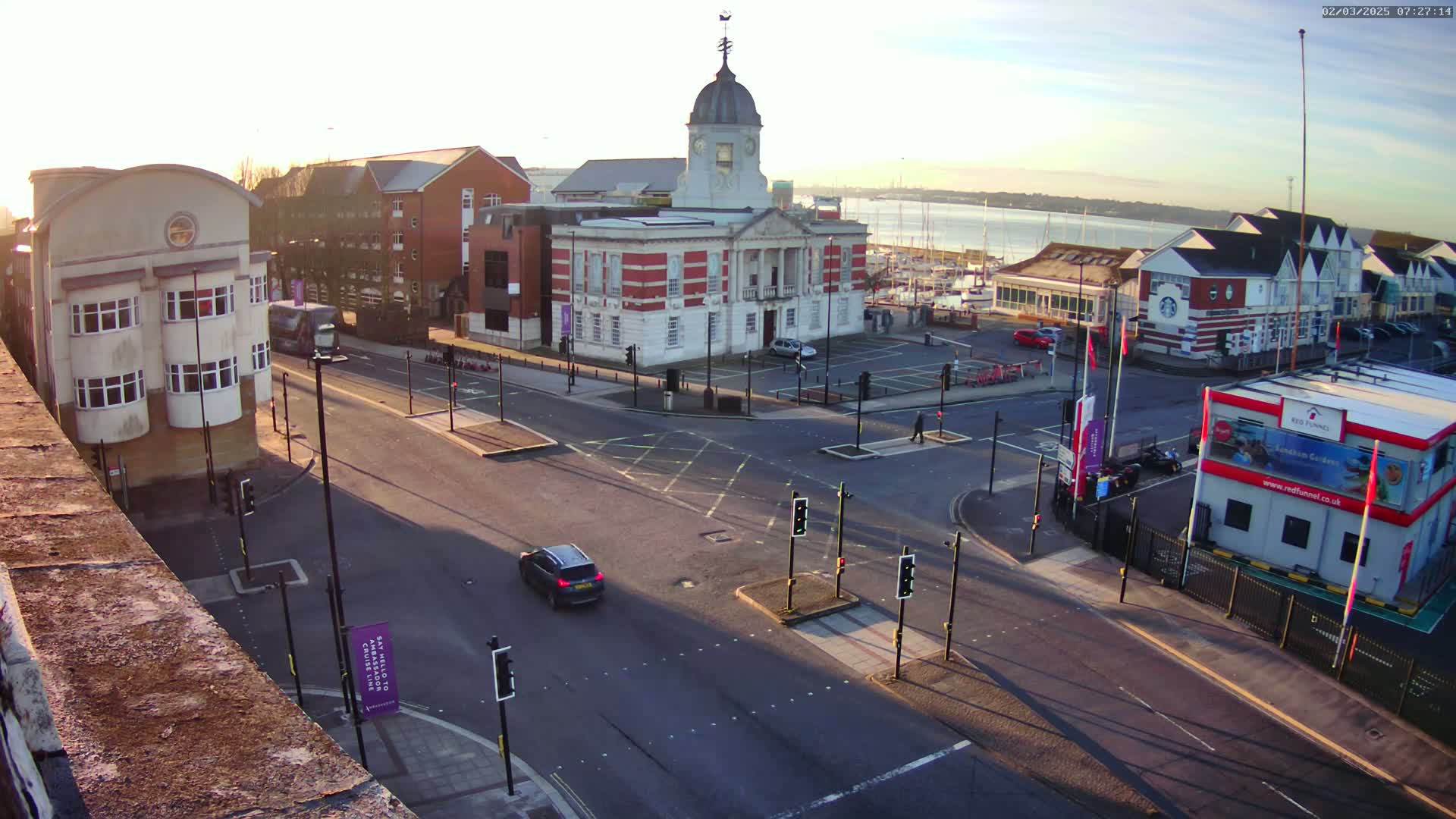 A sunny day reveals a town square with a large brick building at its center, surrounded by streets, cars, and other buildings, overlooking a harbor with boats.