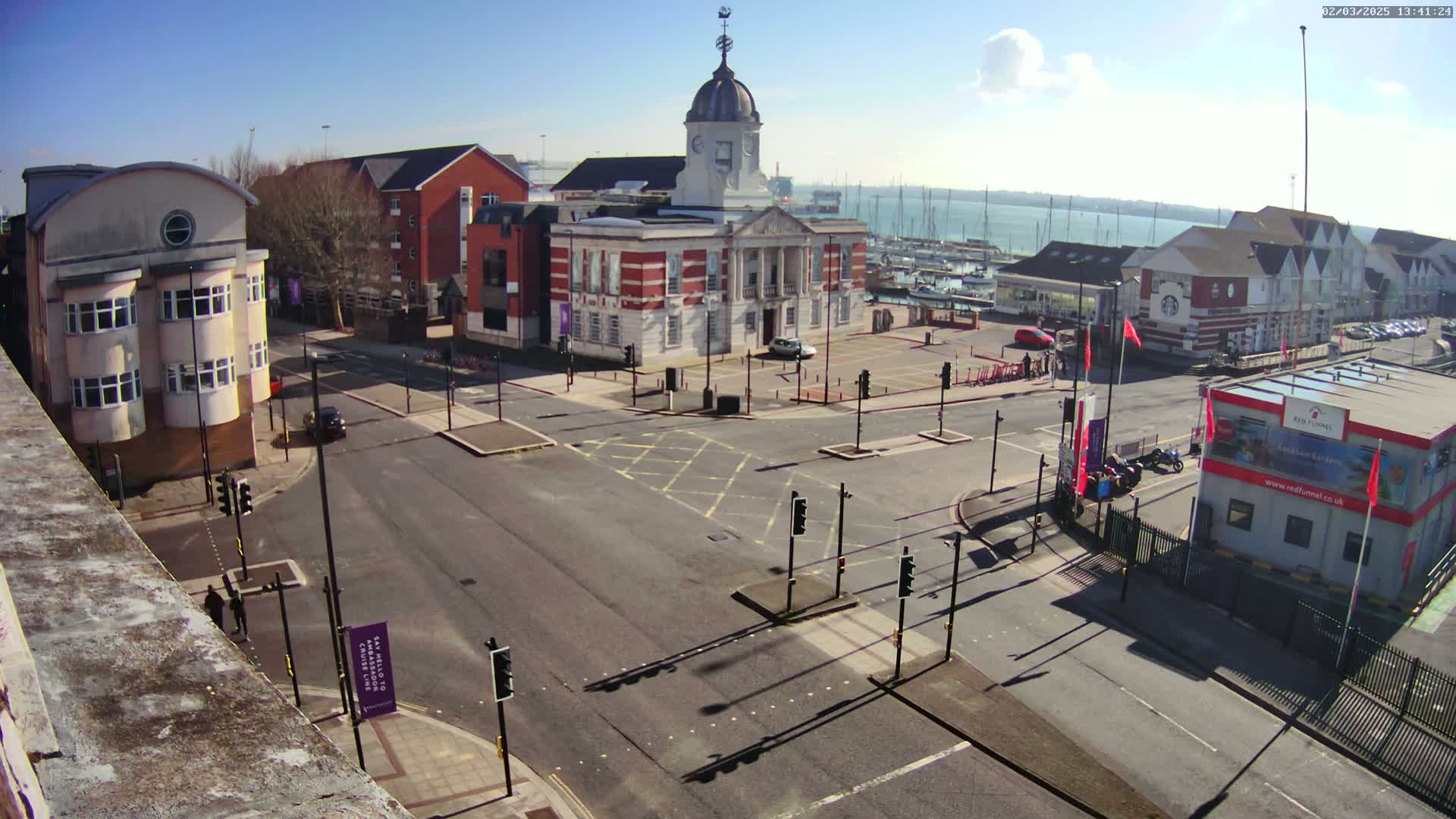 A sunny day reveals a town square with a large brick building, surrounding buildings, cars, and a body of water with boats in the distance.