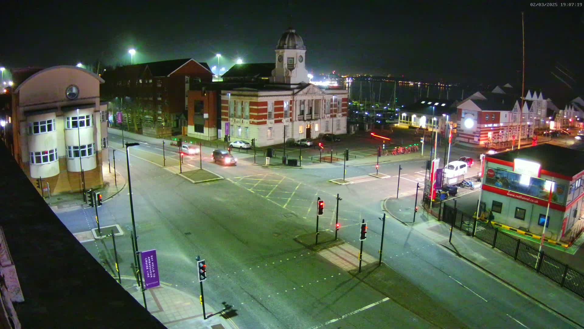 A nighttime aerial view of a city intersection with several buildings, including a large clock tower building, and a few cars and vehicles.