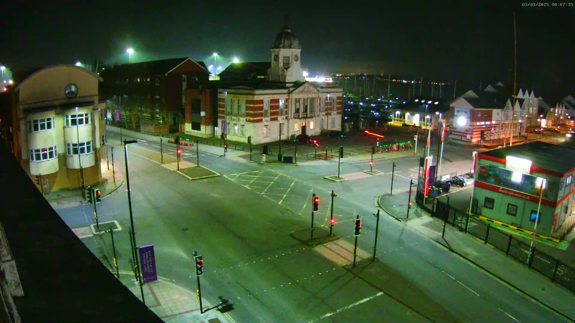 A nighttime, overhead view of a town square with a clock tower building, surrounded by other buildings, roads, traffic lights, and a marina in the background.
