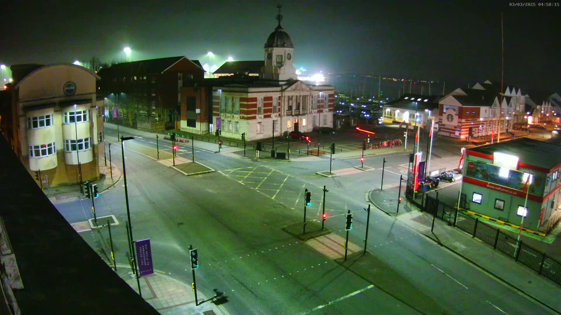 A nighttime aerial view of a street intersection with several buildings, traffic lights, and a few parked cars under foggy conditions.