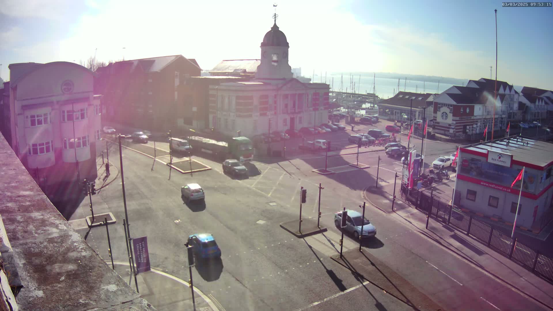 A sunny day reveals a town square with a large building, several cars on the road, and a harbor with boats in the distance.