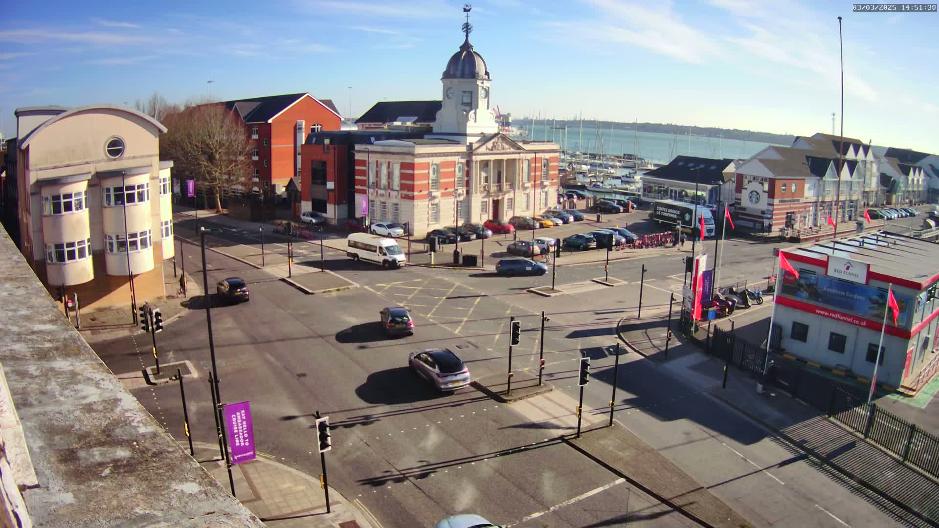 A sunny day reveals a town square with a large brick building, multiple smaller buildings, parked cars, and several cars at an intersection; a body of water with sailboats is visible in the background.