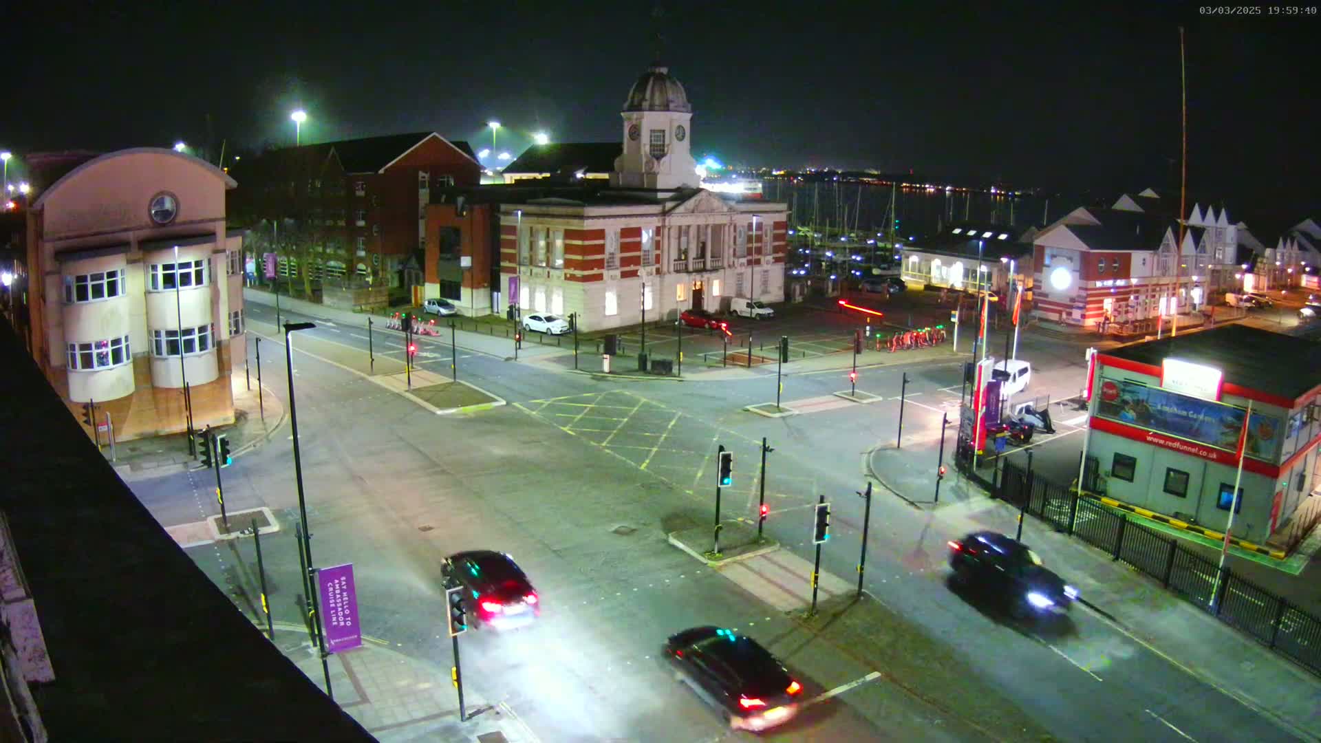 A nighttime aerial view of a town square with a large brick building, several smaller buildings, cars, and traffic lights, under clear skies.