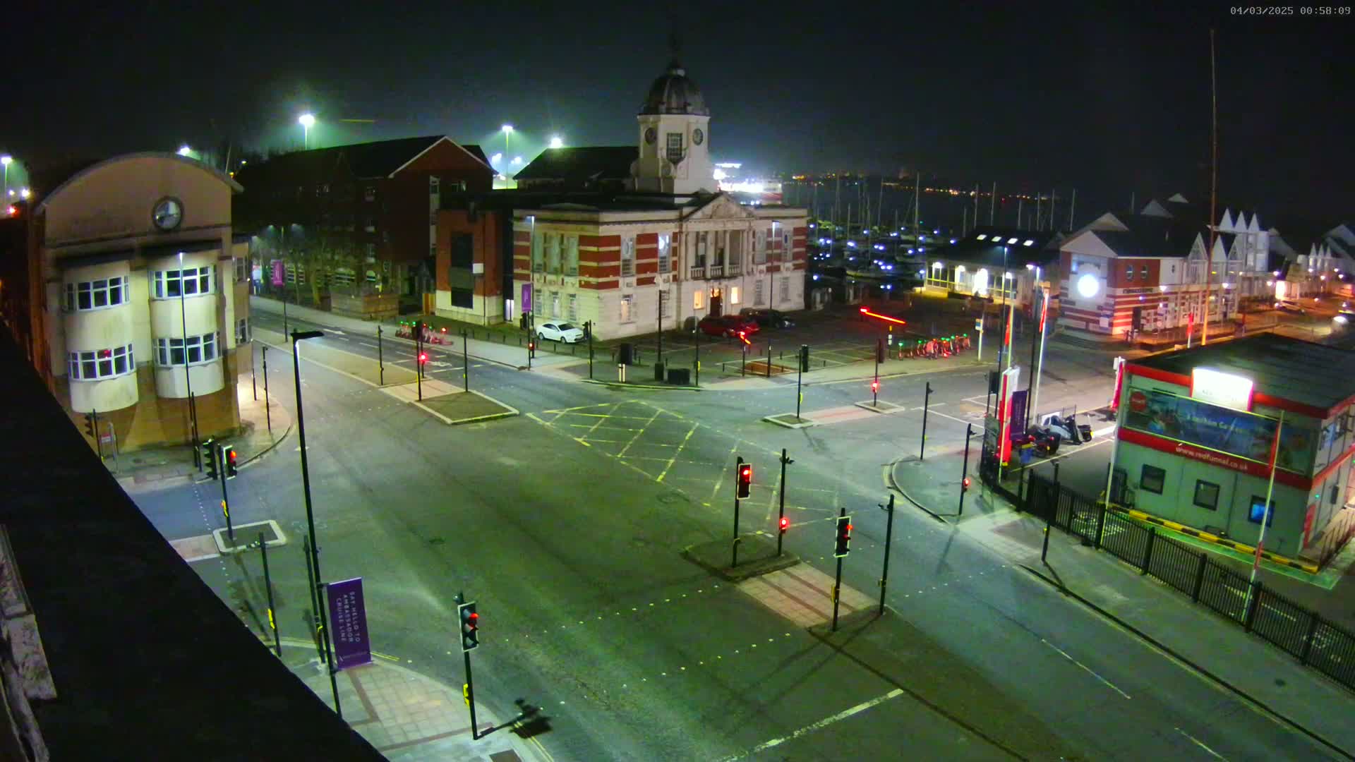 A nighttime, high-angle view of a town square intersection with a clock tower building and a marina in the background under hazy conditions.