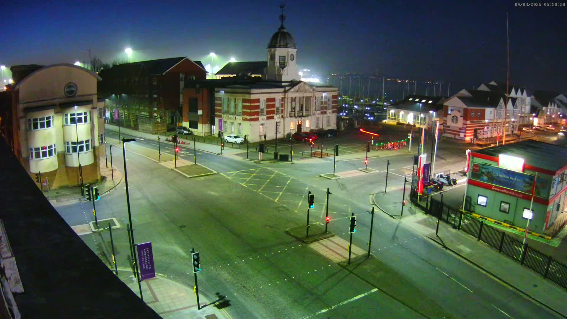 A nighttime aerial view of a town square with a clock tower building and several other structures, under a dark, possibly foggy sky.