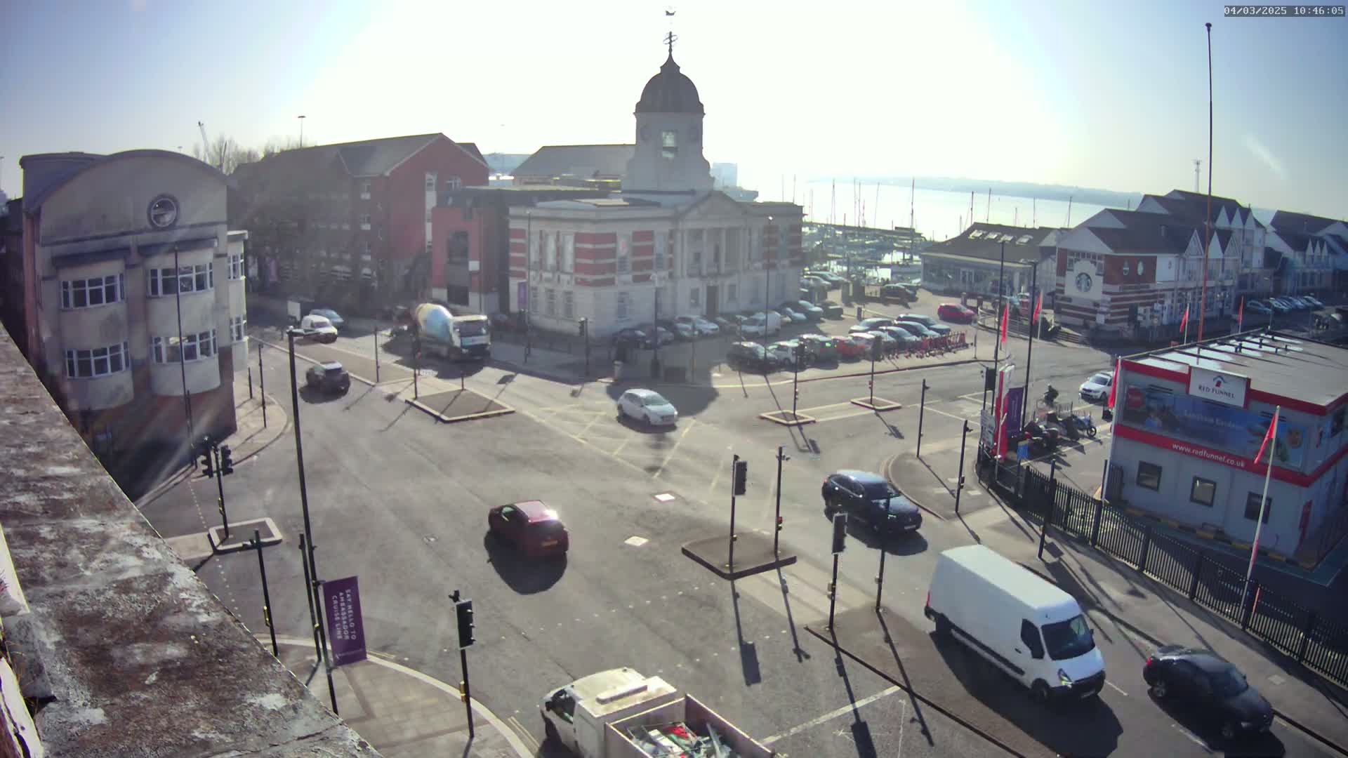 A high-angle view of a town square on a sunny day, showing several cars, a van, and a town hall, with a body of water and sailboats visible in the background.