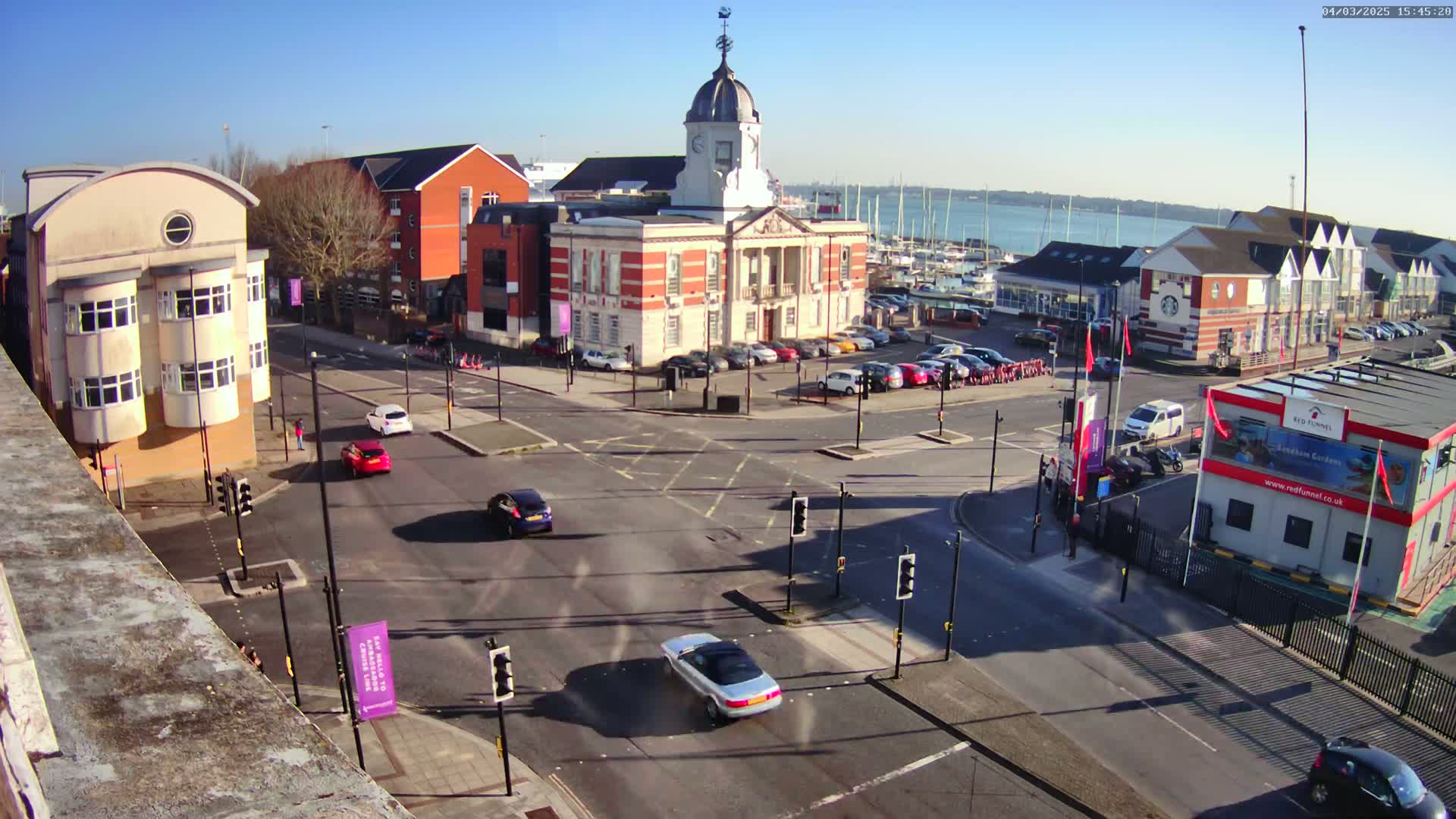 A sunny day reveals a town square with a large brick building, surrounded by other structures, cars, and a marina with numerous boats in the background.