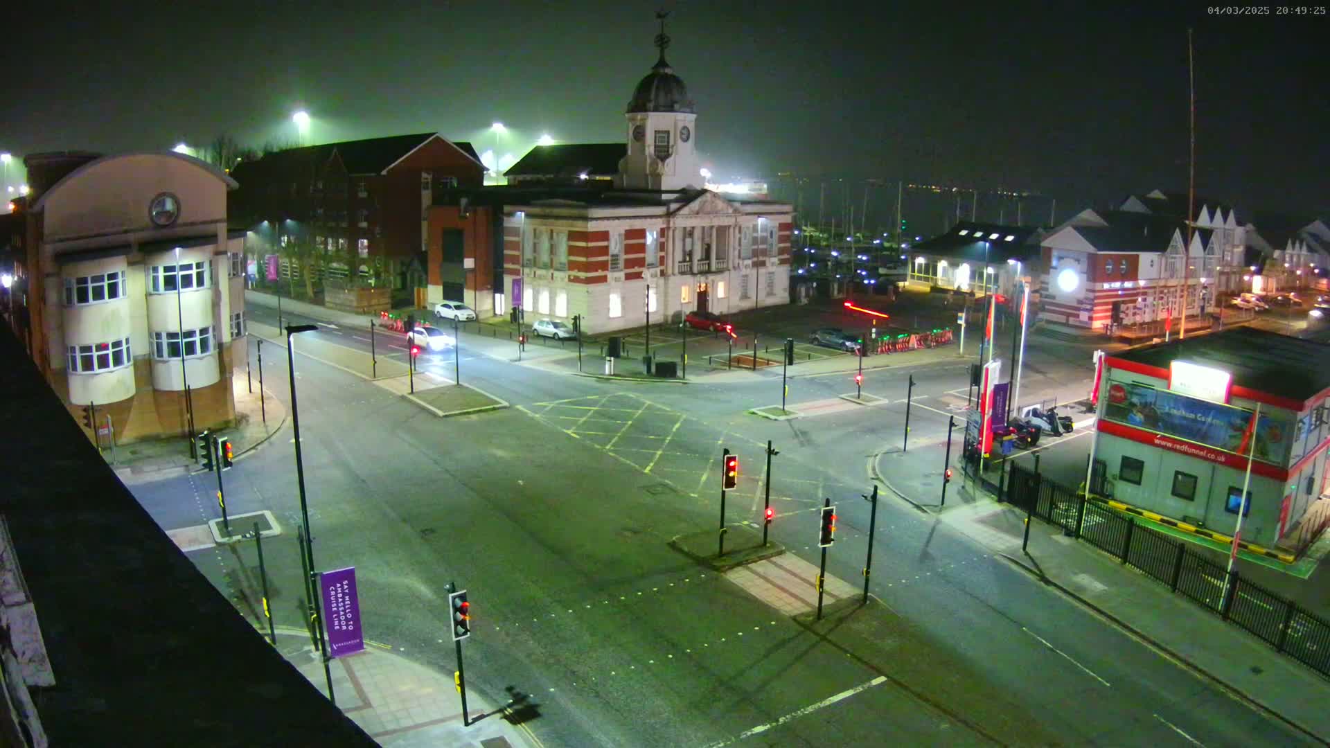 A foggy night scene shows a street intersection with several buildings, including a large, white building with a clock tower, and traffic lights displaying red.