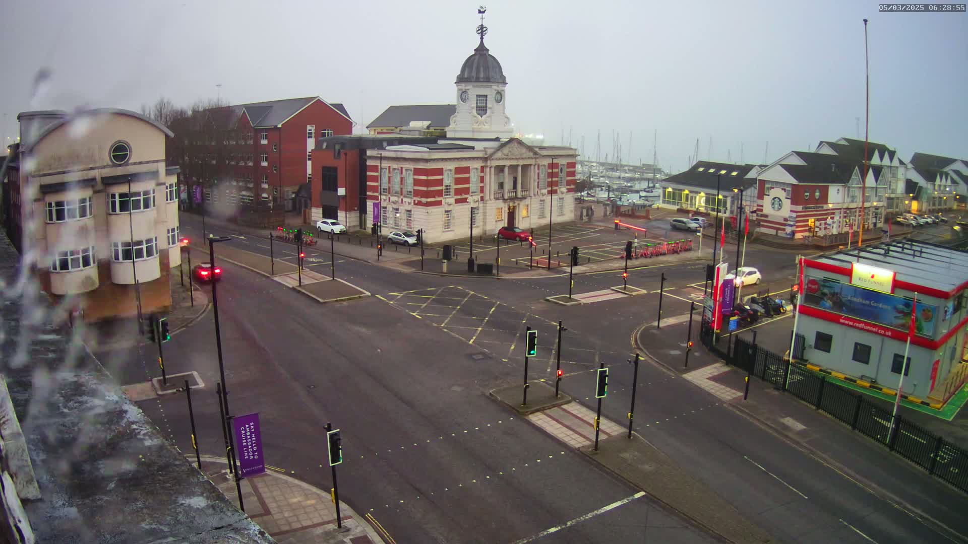 A view of a town square with a large brick building, surrounded by other buildings and a marina in the background, on a cloudy day with light rain or mist.