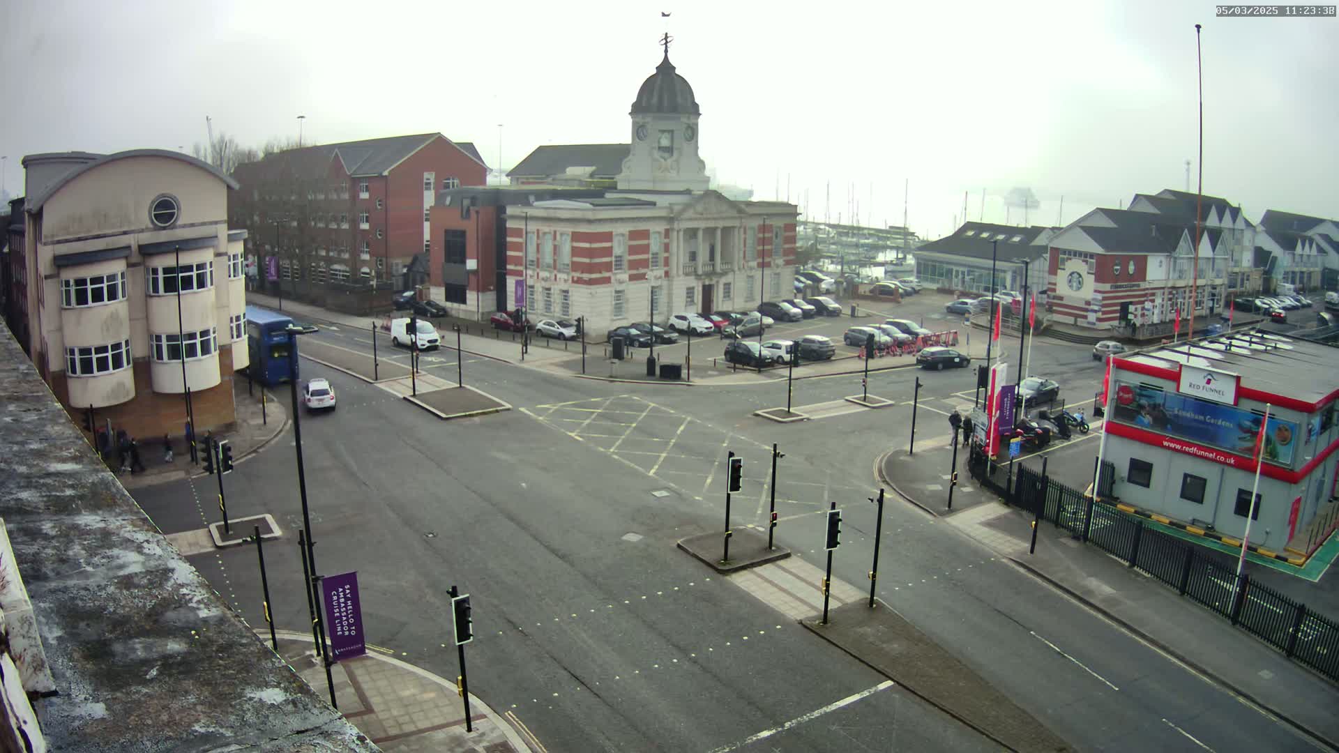 A town square with a brick building, a parking lot, and a road intersection on an overcast day.